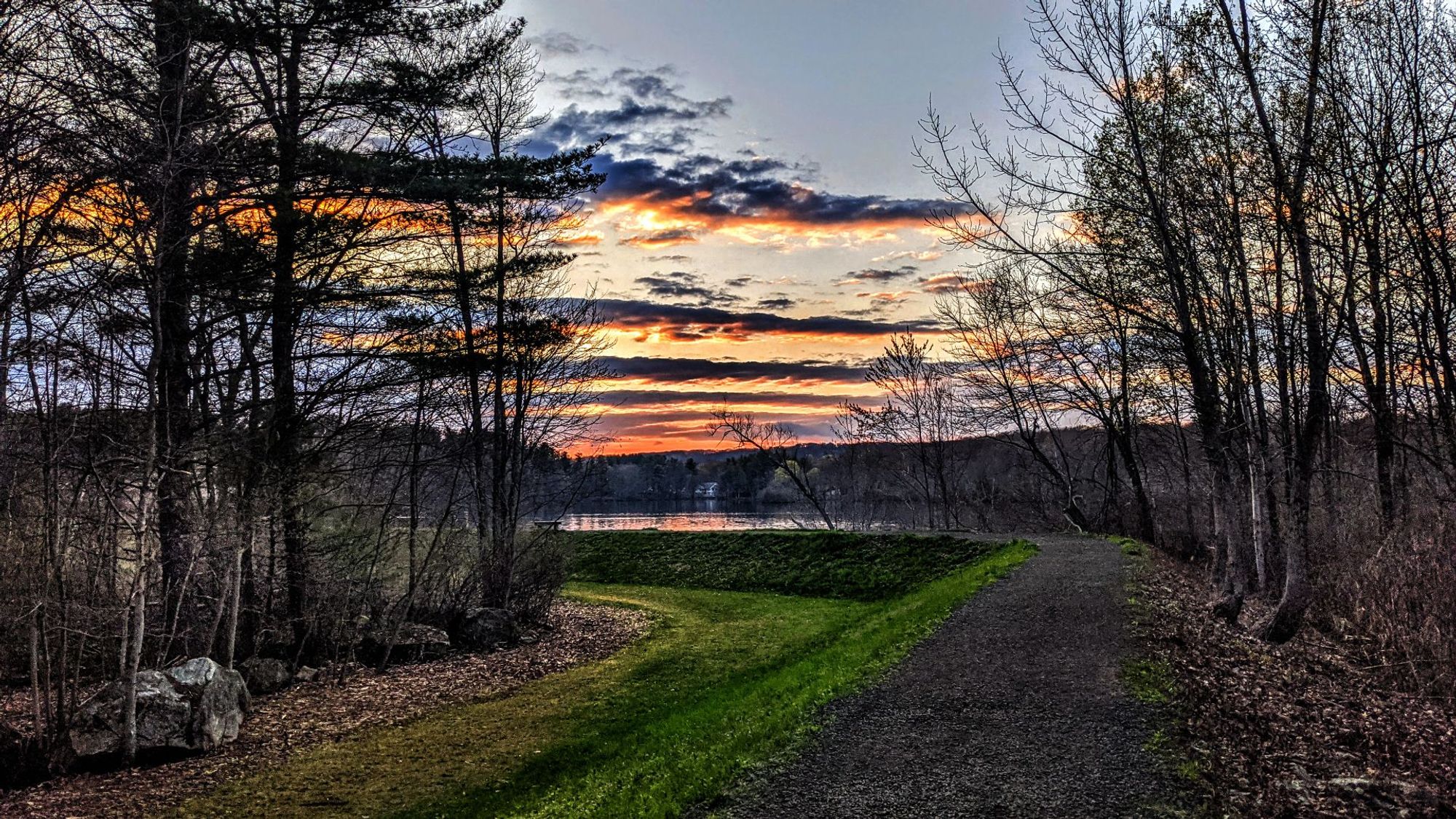 A blue gray evening sky with long, thin gray clouds tinted orange and gold by the setting sun. A wide dirt and gravel path follows the top of an embankment rising several feet above the surrounding ground. The left side of the path has a wide swath of bright green spring grass running down the left slope of the embankment and a few more feet across the level ground below. To the right of the path and to the left of the grassy area are forests of leafless hardwoods beginning to bud with a hemlock or two mixed in. The path jogs sharply left not far ahead as it comes to the shore of a lake. A similar forest, with a few houses mixed in, covers the far shore of the lake. A wooden bench is on our shore near the point where the path disappears behind the trees on our left.