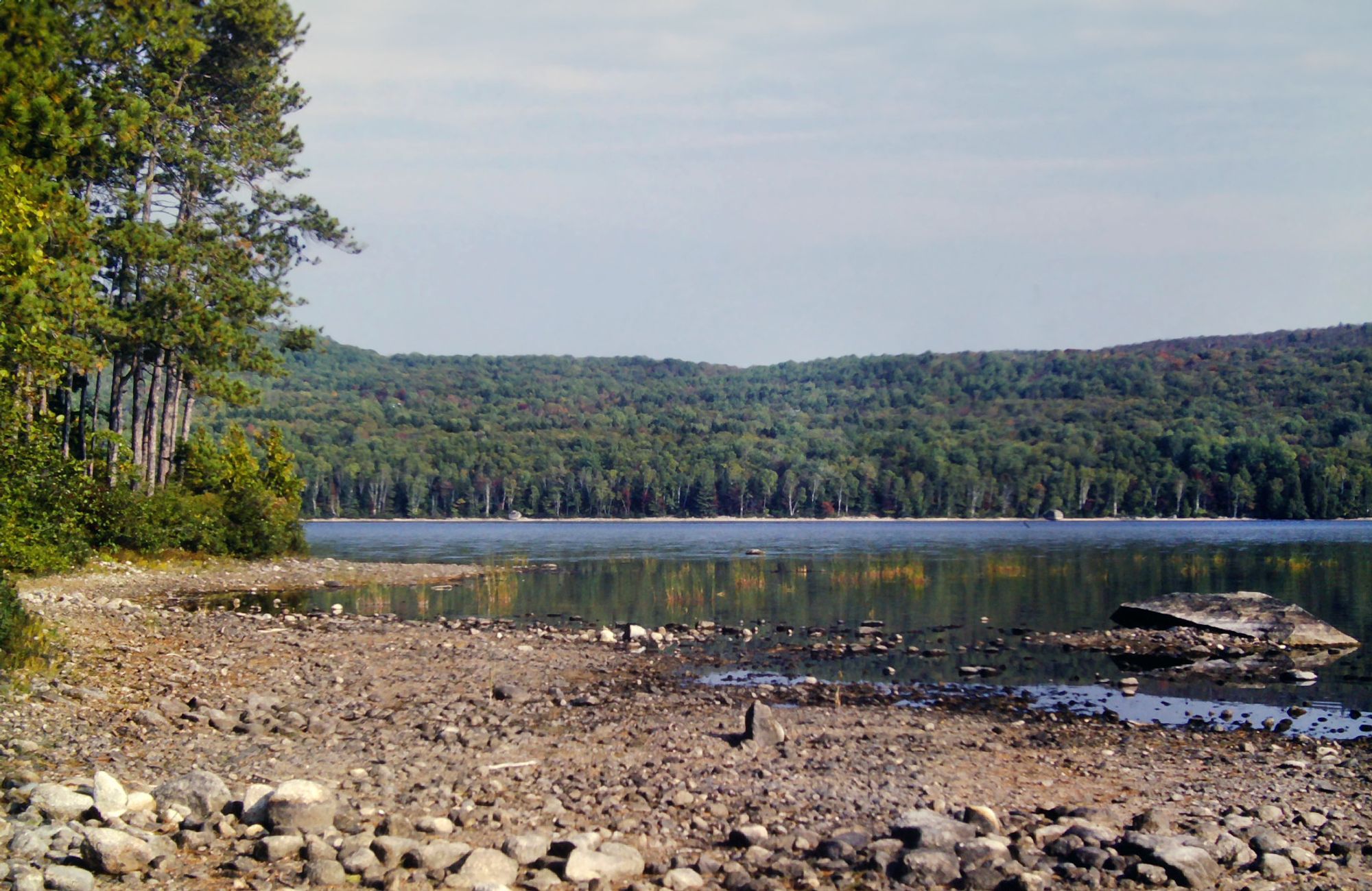 A hazy, soft blue sky with gauzy, streaky clouds. A wooded peninsula on the shore of a large lake. A long drought has exposed a great deal of rock and gravel covered lakebed. A low mountain rises above the far shore, its slopes are covered with a predominantly evergreen forest. A few hardwoods are showing autumn golds and oranges. A breeze is barely rippling the clear lake waters.