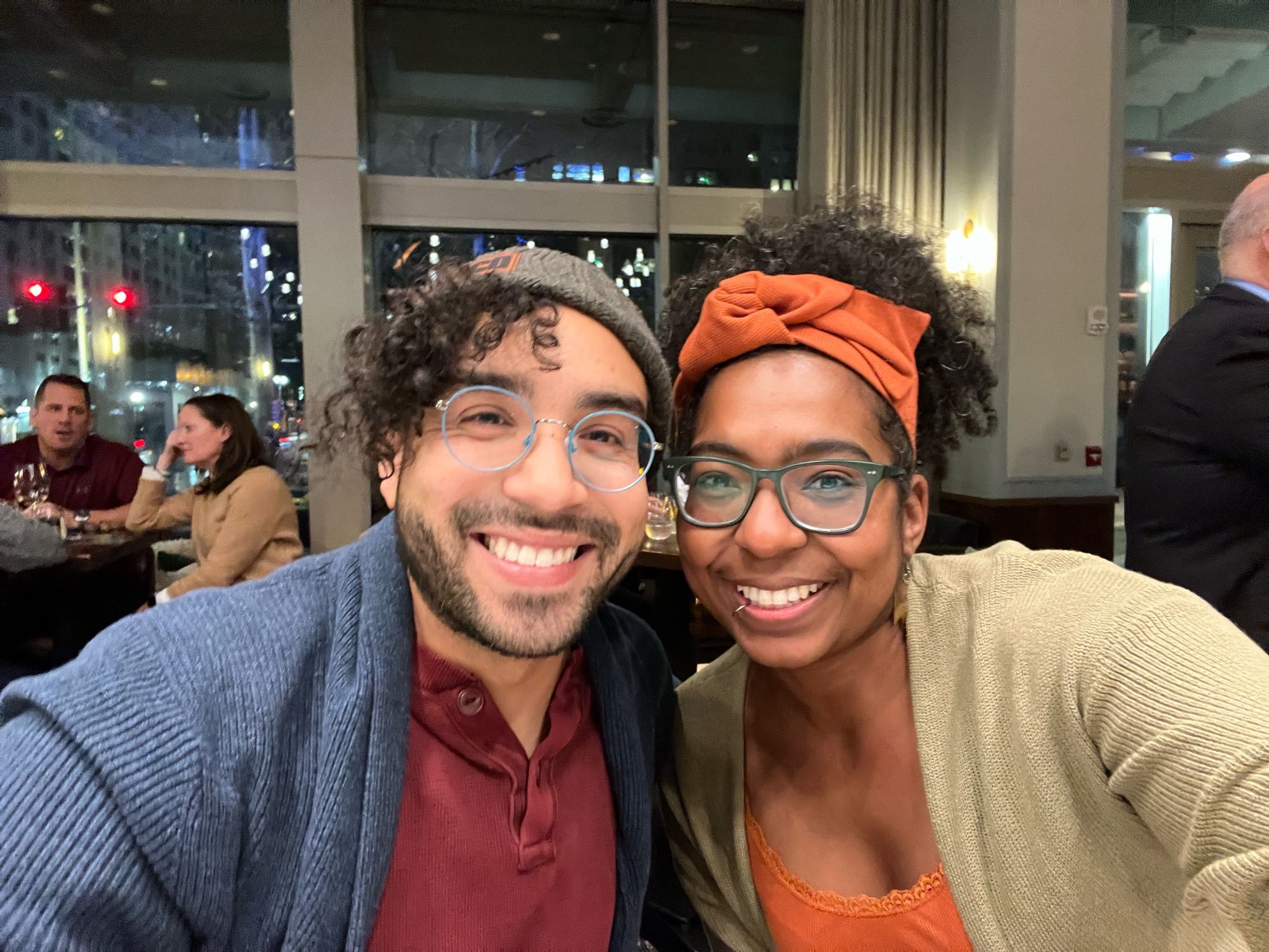 Photo of two friends smiling at the camera. Both are black wearing teal/green glasses, have curly hair, and are wearing sweaters. On the left is Ed, a man, and the right, me, a woman.