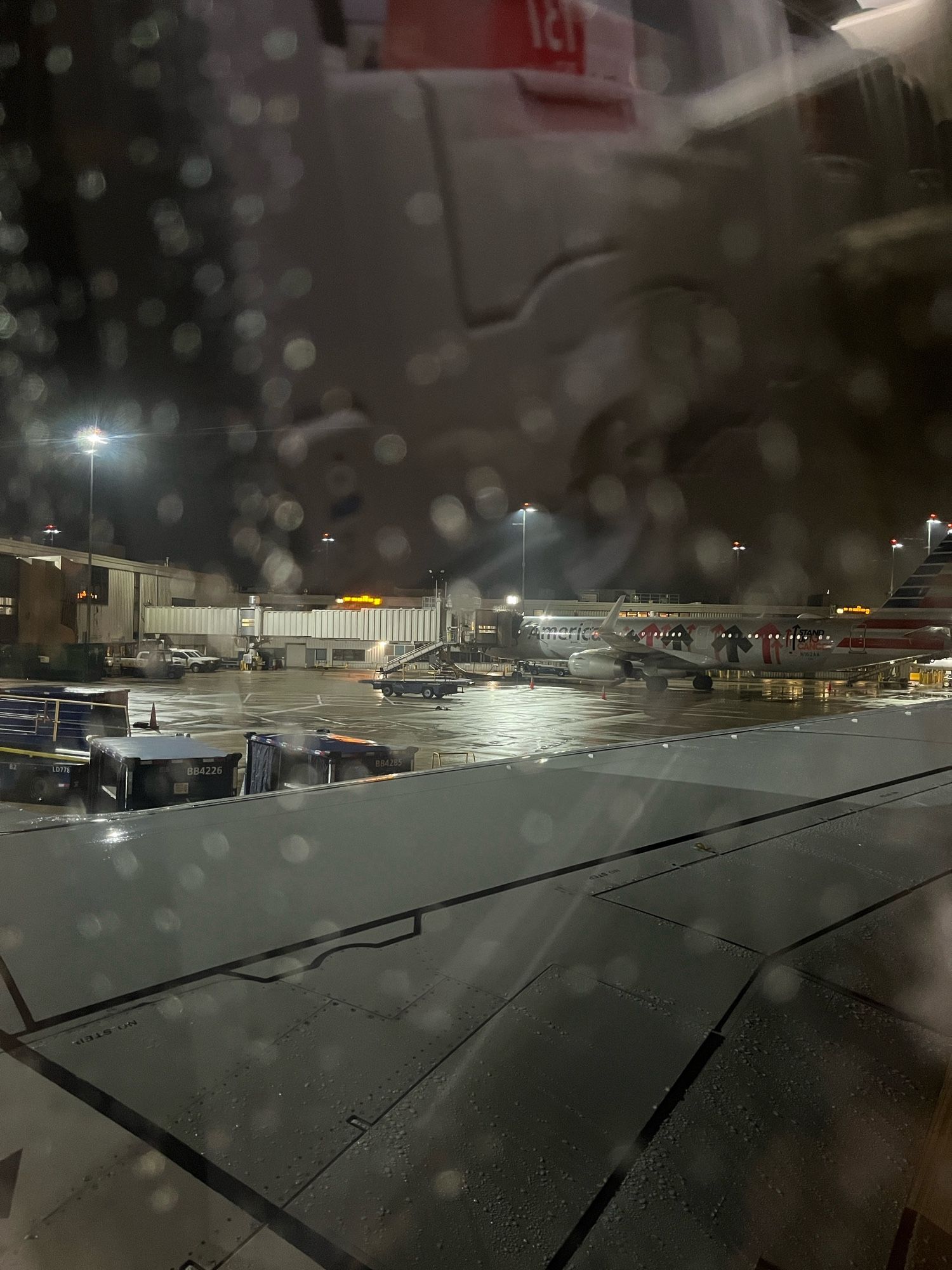 Photo looking out from a grounded airplane window. There are out of focus raindrops on the window glass, the plane wing takes up a large percentage of the photo and in the top third is the tarmac with another plane with boarding bridge attached.