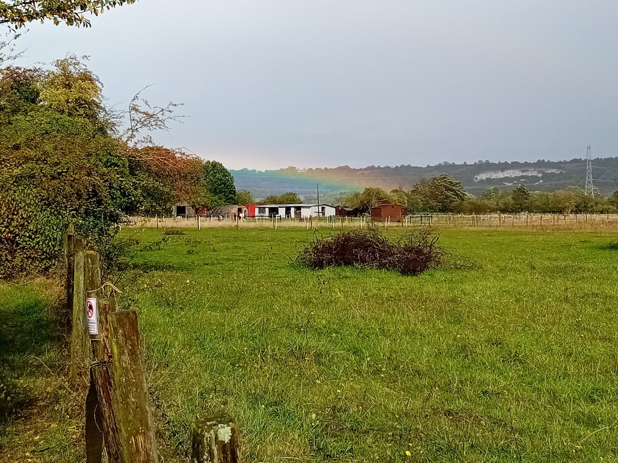 View across a field towards a low rainbow, just above a farm building.