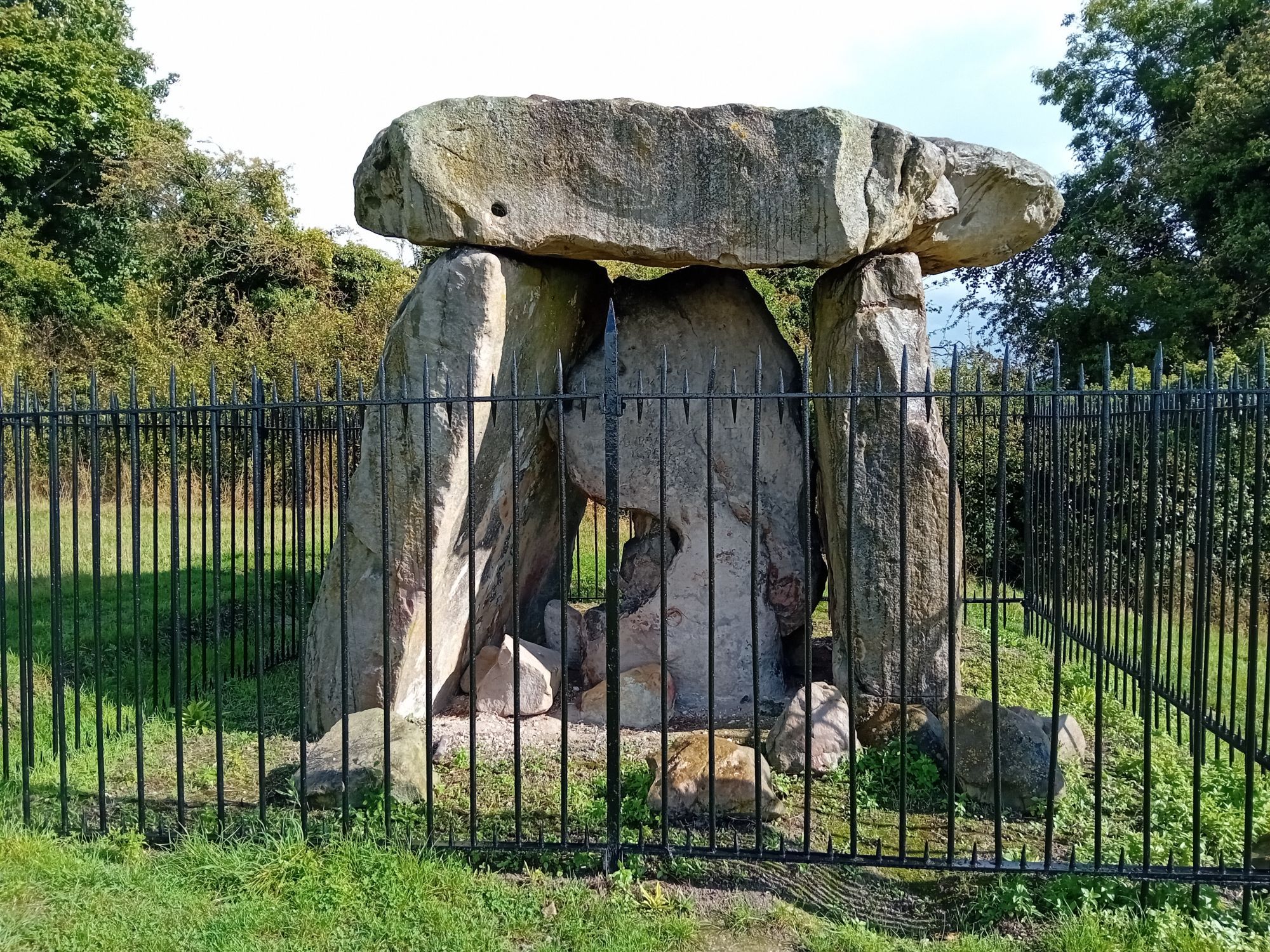 A stone megalith. There are two upright stones, with a flat stone across the top. There is another stone in the middle. The megalith is behind a metal fence.