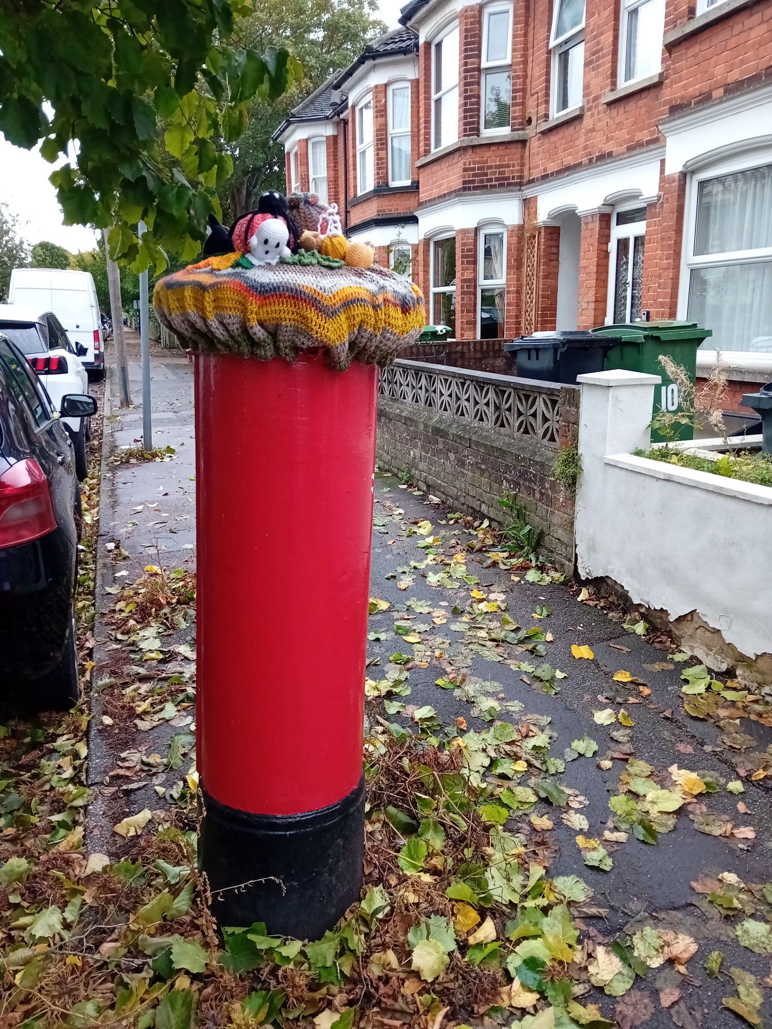 A red pillar box with a crocheted topper on top. The base of the cover is orange. There are crocheted ghosts, spider, cobweb, and squash on top.