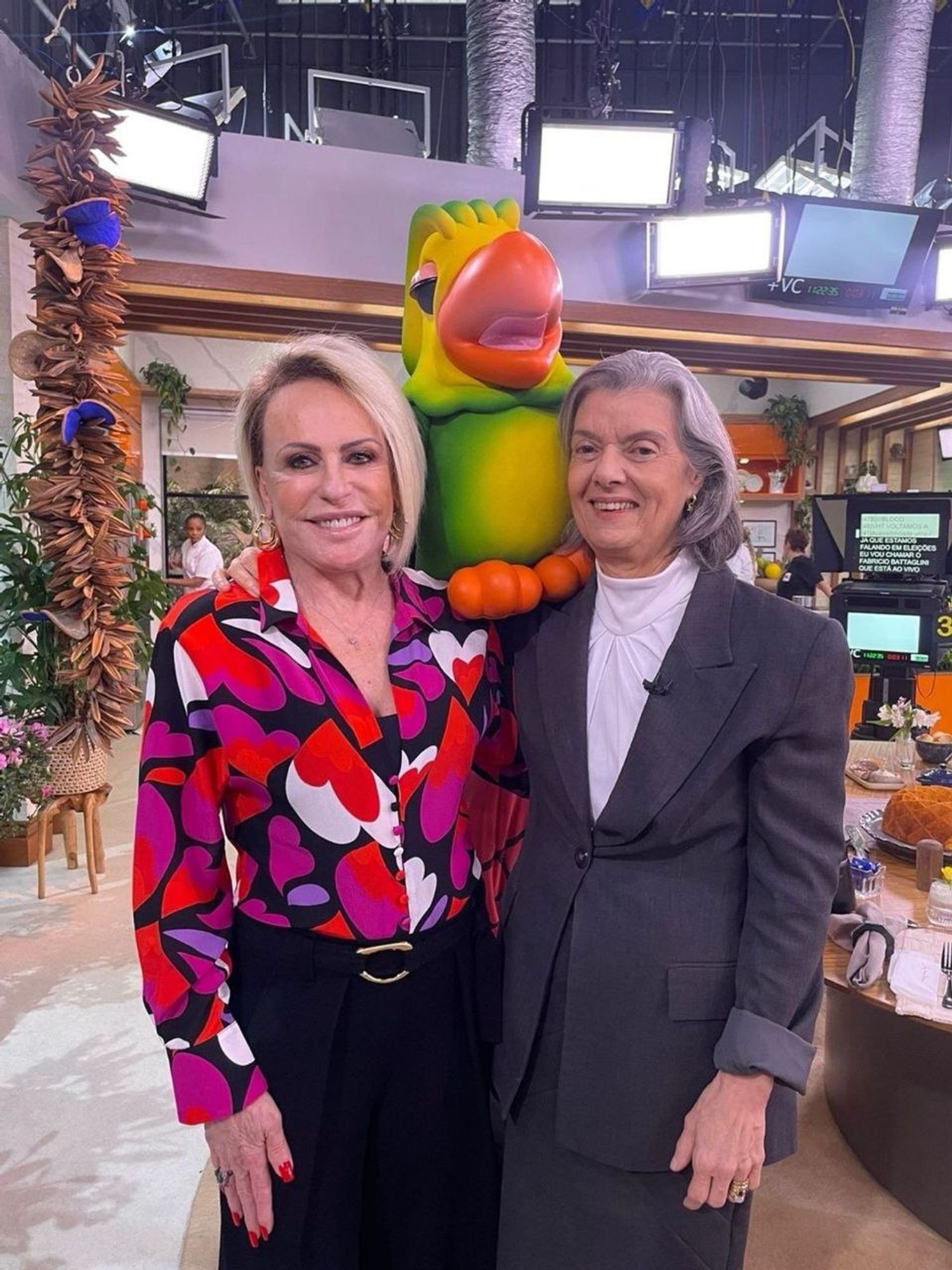 À esquerda, Ana Maria Braga, apresentadora de TV, veste uma blusa colorida em tons de vermelho, roxo e branco com estampas abstratas e calça preta. À direita, está a ministra do STF Cármen Lúcia, usando um terno cinza escuro com uma blusa branca. Entre elas, há uma grande réplica do personagem Louro José, um papagaio com penas verdes e um grande bico laranja. Ao fundo, é possível ver parte do cenário de um programa de TV, com luzes de estúdio, plantas decorativas e uma mesa com alimentos.