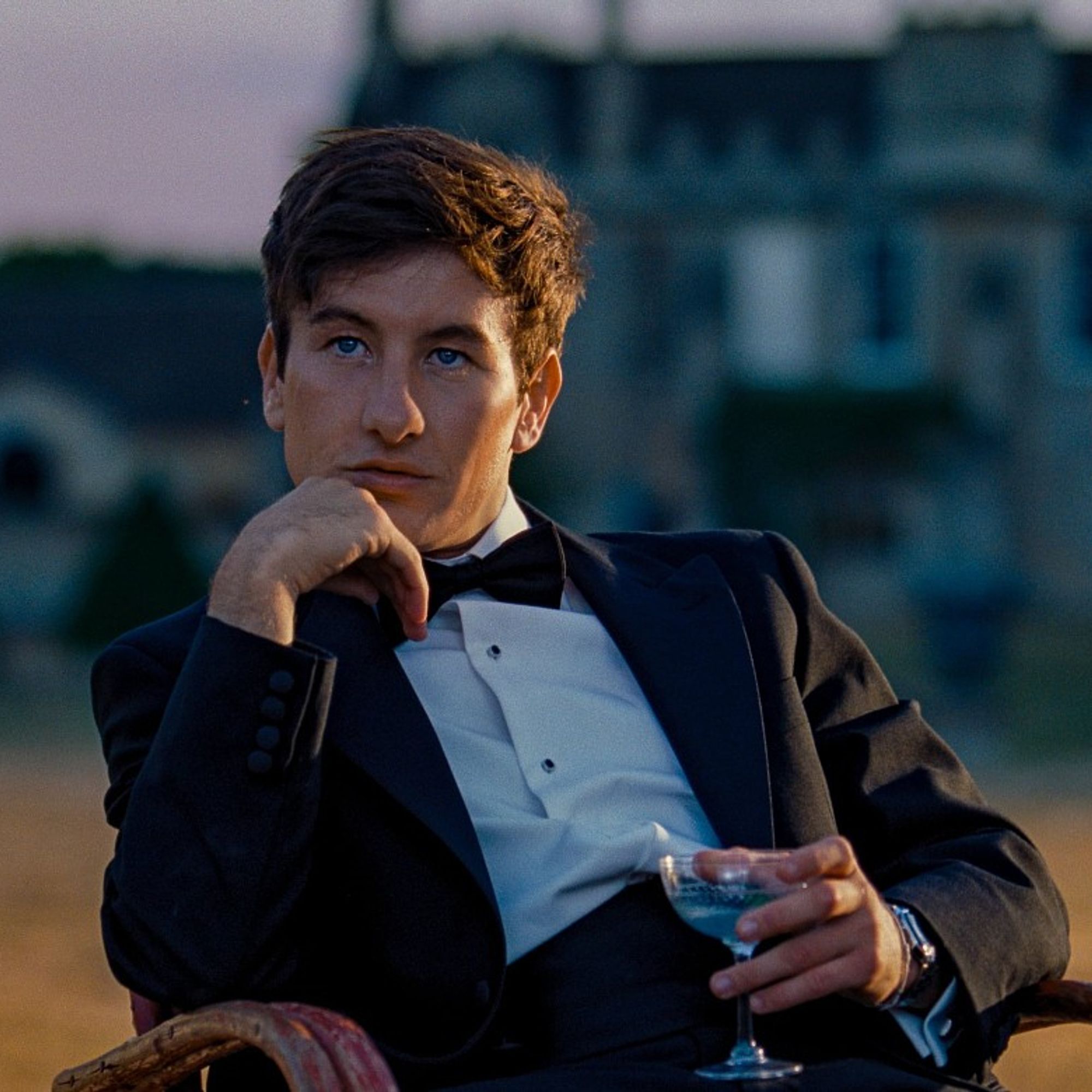 A imagem mostra Barry Keoghan no papel de Oliver Quick no filme Saltburn. Ele está sentado em uma cadeira ao ar livre, em frente a um cenário que parece ser uma grande mansão ao fundo, desfocada. Keoghan veste um elegante smoking preto com gravata borboleta e camisa branca. Ele segura um copo de coquetel em uma mão e apoia o queixo na outra, com uma expressão pensativa e distante. A luz suave do entardecer ilumina a cena, criando uma atmosfera sofisticada e contemplativa.