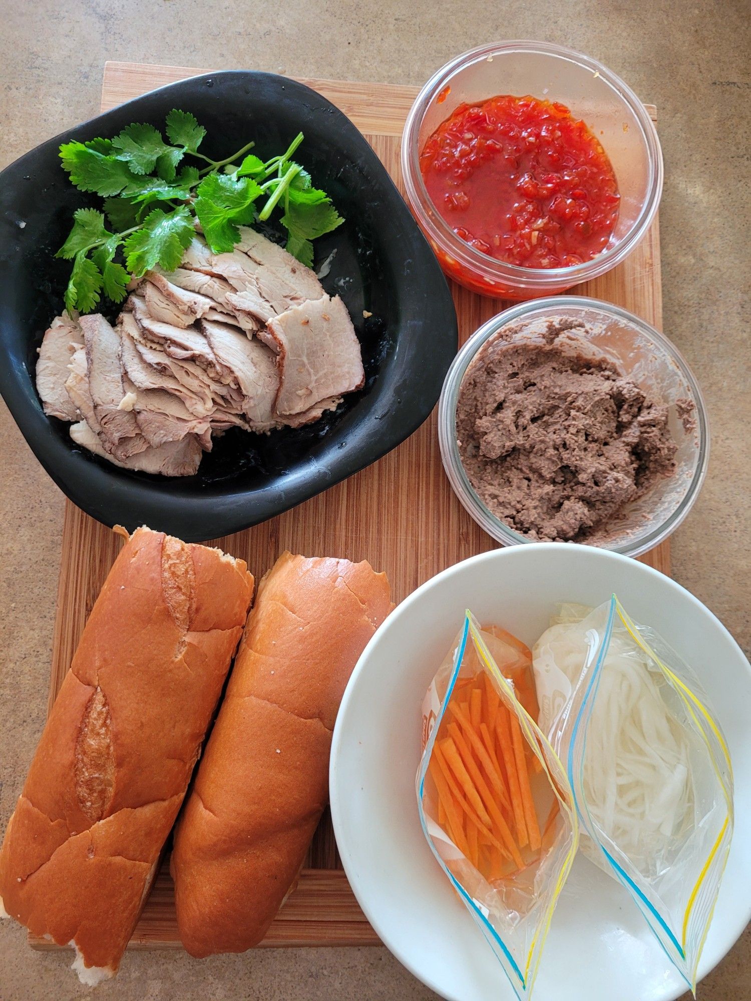 Ingredients clockwise from top left of a black soup plate with sliced roast pork & sprigs of coriander, a small bowl of chilli jam, a small bowl of homemade pork liver & mushroom pate, a bowl with pickled daikon & pickled carrot in Ziploc bags, two bread rolls.