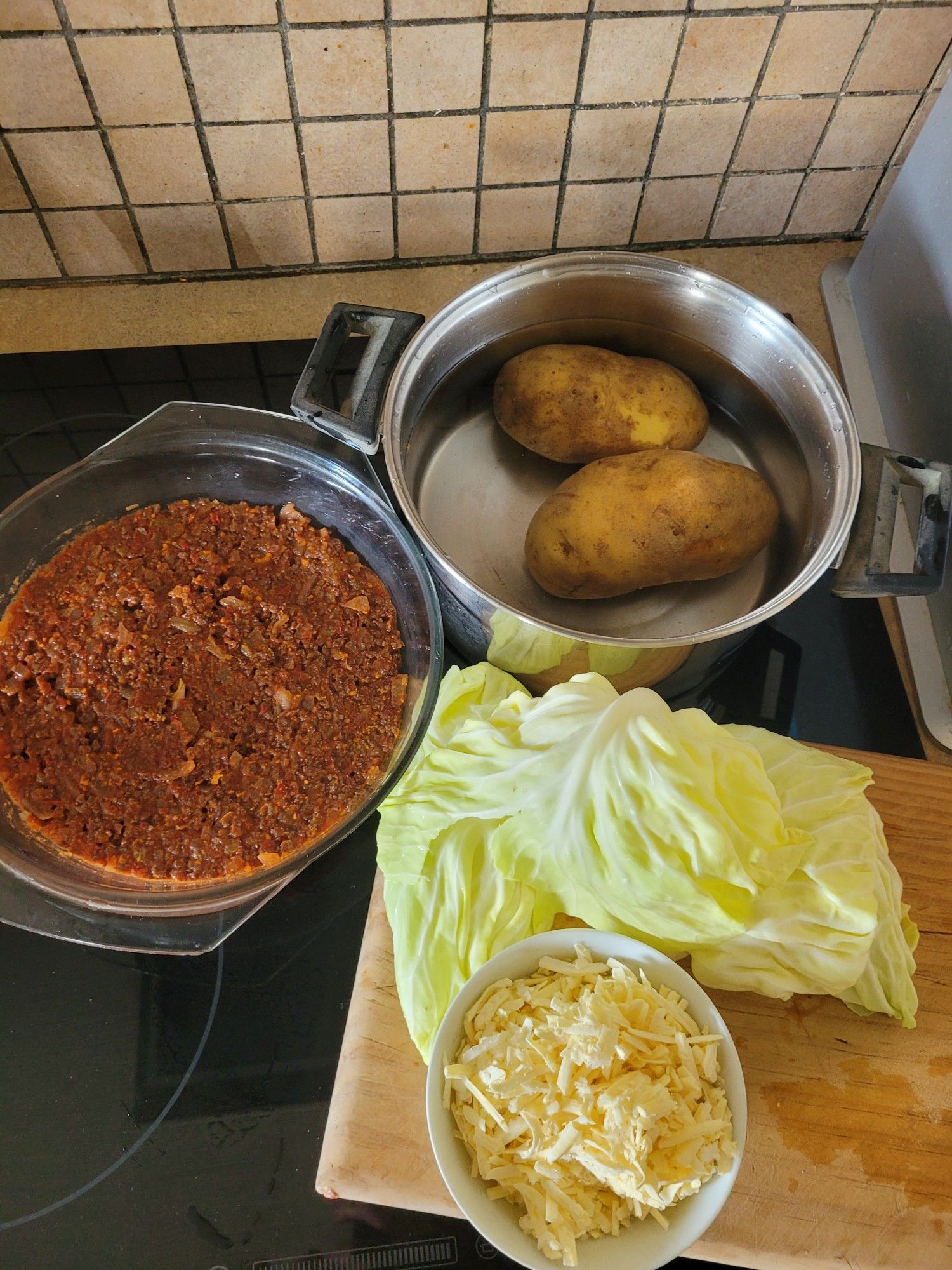 Ingredients clockwise from left of a casserole dish with cooked beef mince, a pot of water with potatoes, a chopping board with cabbage pieces & a small bowl of grated cheese.