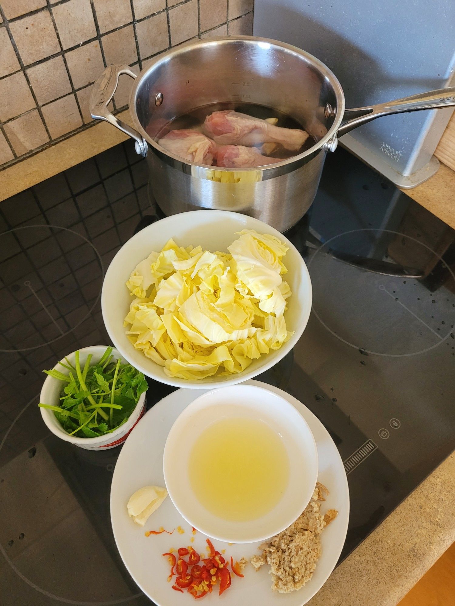 Ingredients from top of a pot with chicken drumsticks in water, a bowl of cabbage pieces, a ramekin with chopped coriander, a round white plate with a small bowl of lemon juice, a clove of garlic, minced ginger, chopped red chilli.