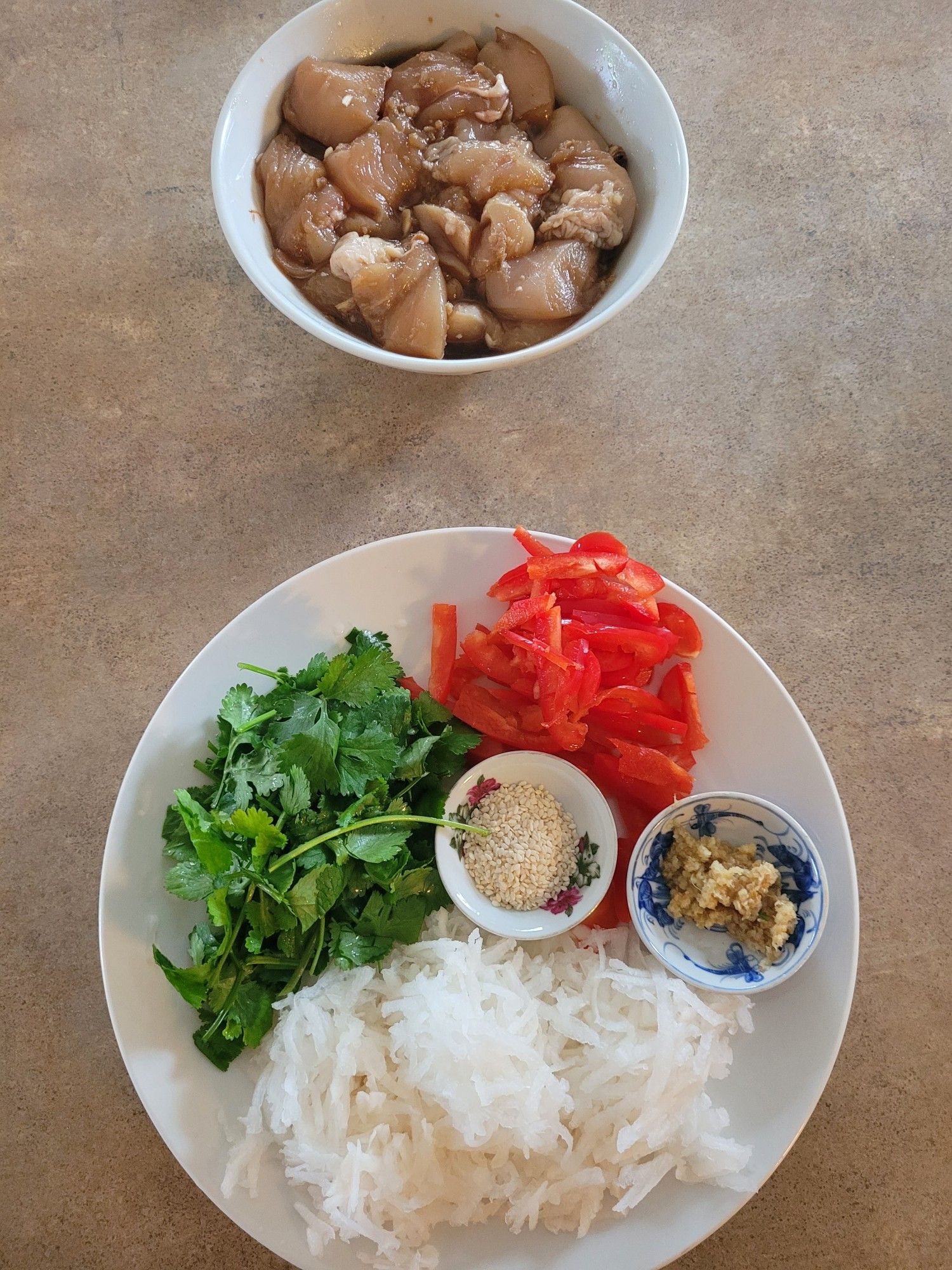 Ingredients at top of a bowl of marinated chicken breast pieces & at bottom a round white plate with thin sliced red capsicum roughly chopped fresh coriander, grated daikon, and two small dishes with grated ginger & sesame seeds respectively.