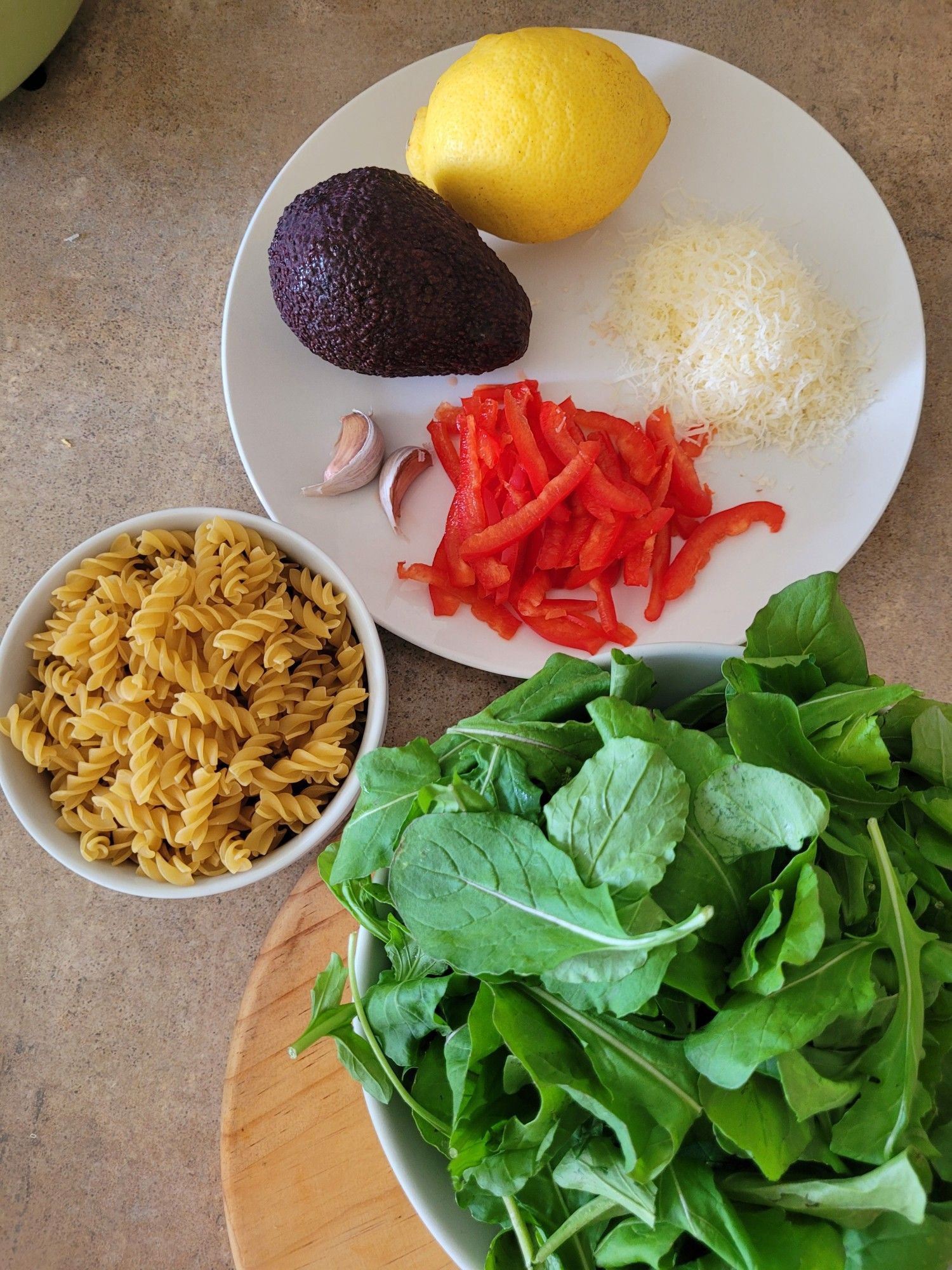 Ingredients from top of a round white plate with an avocado, a lemon, two cloves of garlic, red capsicum cut into strips, grated cheese. A small bowl of dry pasta spirals, a bowl of baby rocket leaves.