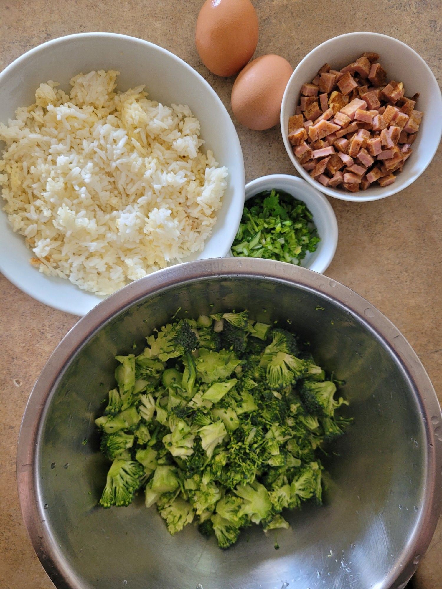 Ingredients clockwise from top left of a bowl of cooked rice, two eggs, a small bowl of diced spam, a ramekin of chopped coriander, a bowl of broccoli pieces.