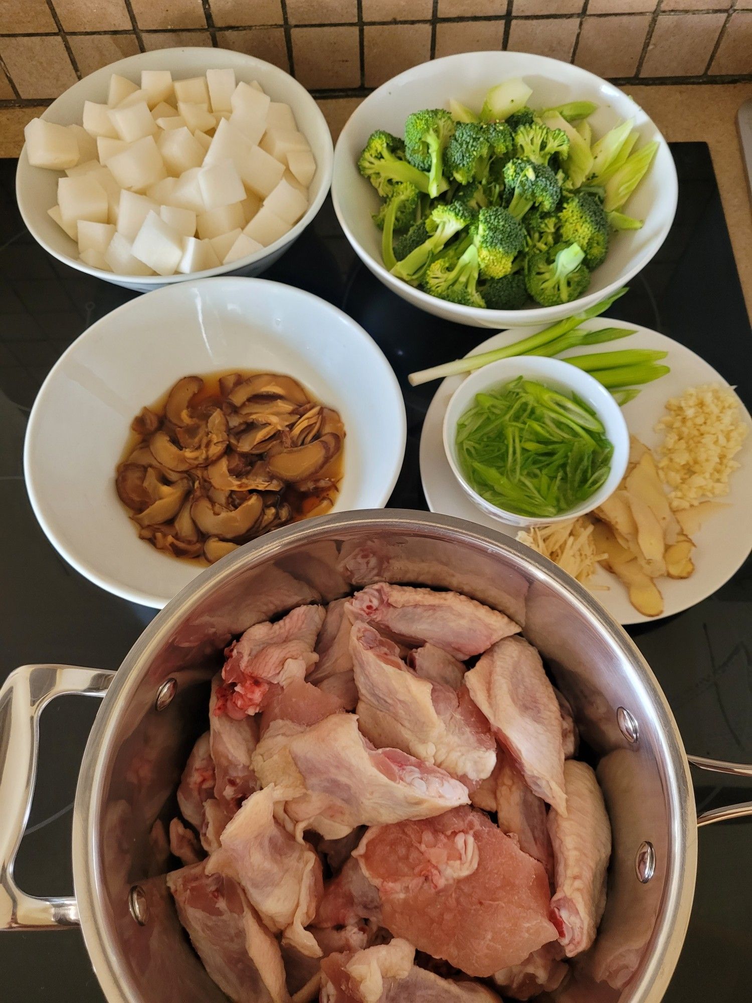 Ingredients clockwise from top left of a bowl of daikon cubed, a bowl of broccoli pieces, a plate with spring onion, sliced ginger, minced garlic, a pot with chicken wing pieces, a bowl with thinly sliced shiitake mushroom