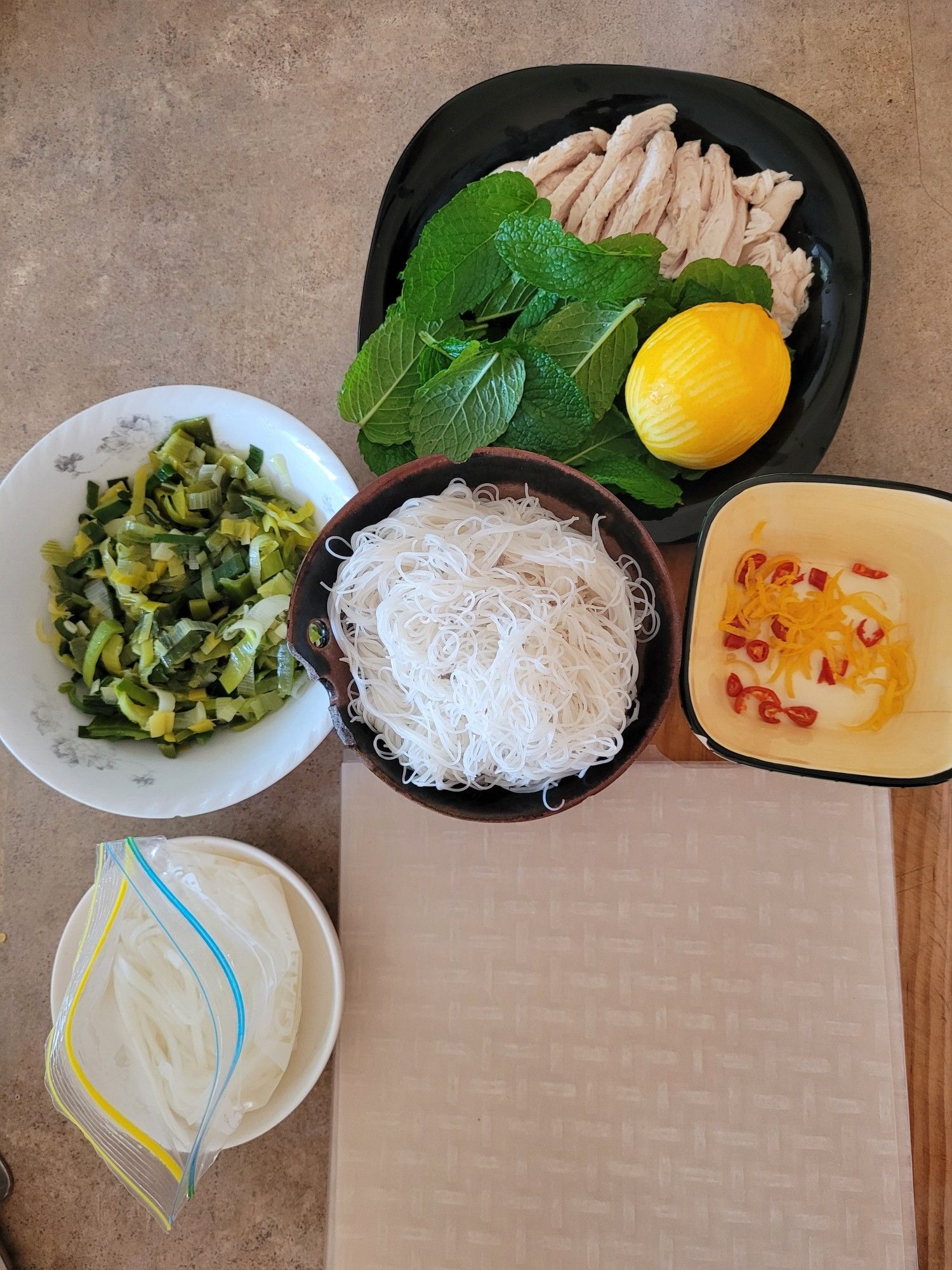 Ingredients clockwise from left of a bowl of leftover sauteed leek, a black soup plate with strips of poached chicken meat, mint leaves, a whole lemon, a square ramekin with chopped red chilli & lemon zest, a bowl of cooked rice vermicelli, a wooden chopping board with square sheets of rice paper, a small bowl of pickled daikon strips.