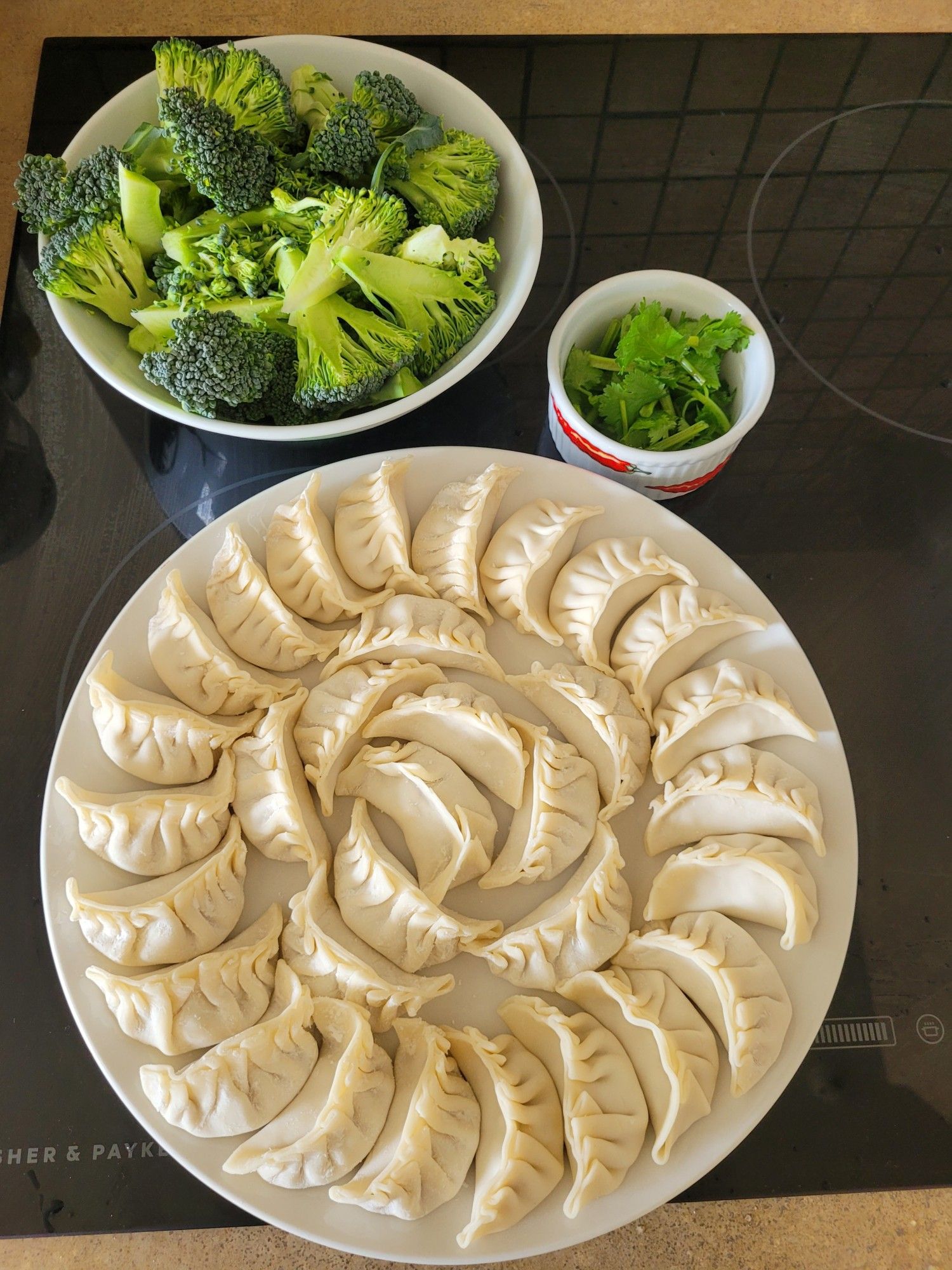 Dinner ingredients from top of a bowl of broccoli pieces, a ramekin with roughly chopped coriander, a round white plate with homemade dumplings.