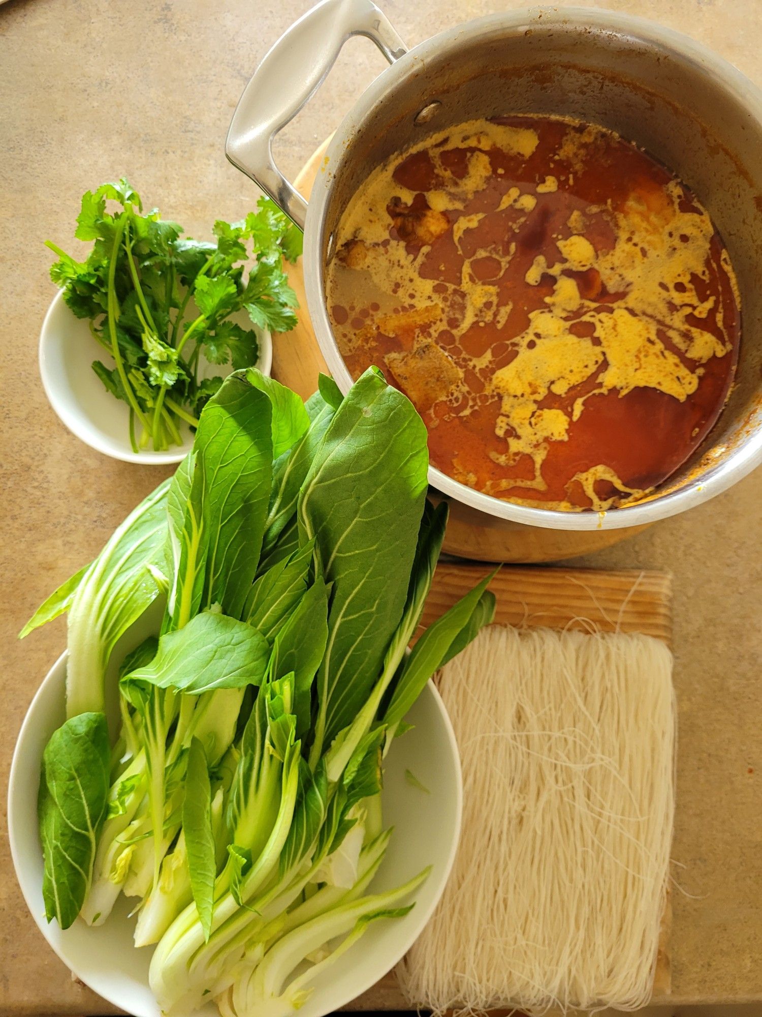 Ingredients clockwise from top left of a bowl of coriander, a pot of chicken and potato curry, a wooden chopping board with rice vermicelli, a bowl of bokchoy sliced lengthways.