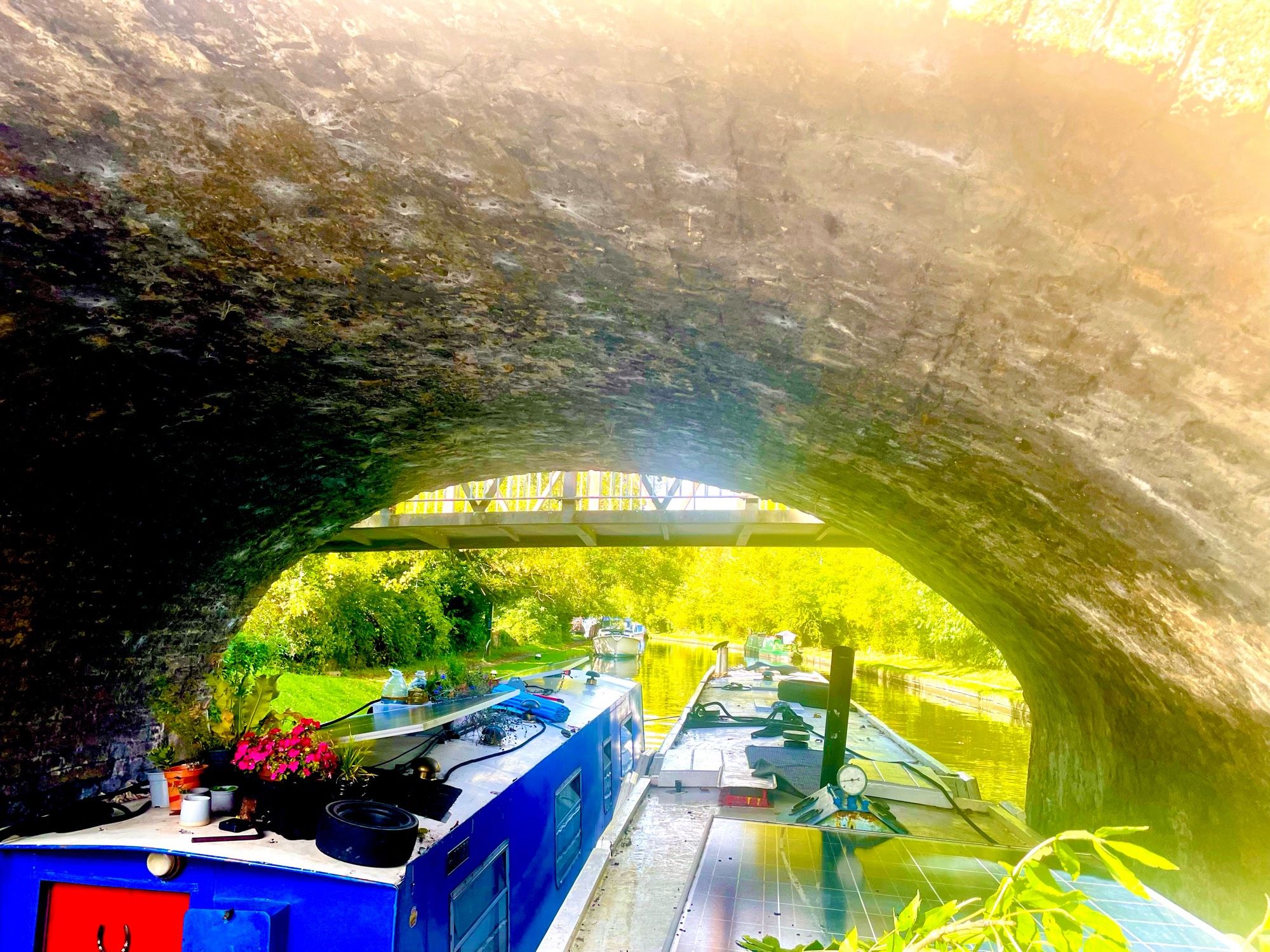 A pair of narrowboats, breasted up, squeezing under a brick arched road bridge, in the distance is the waterway with moored boats on both sides. The towpath is on the right. Above the arch of the bridge the base of a steel footbridge is visible.