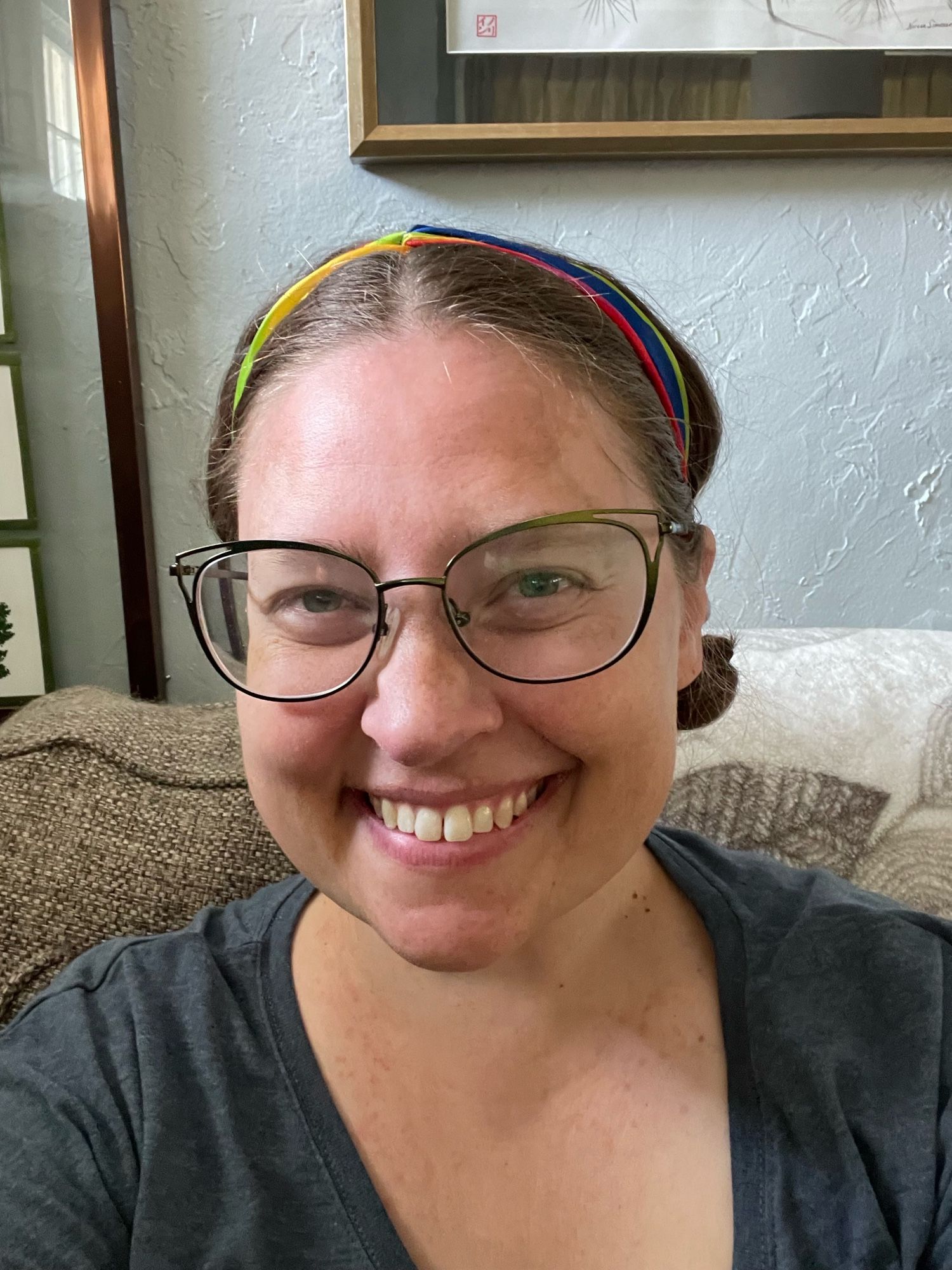 A middle aged white woman smiles at the camera. She has glasses and a rainbow headband.