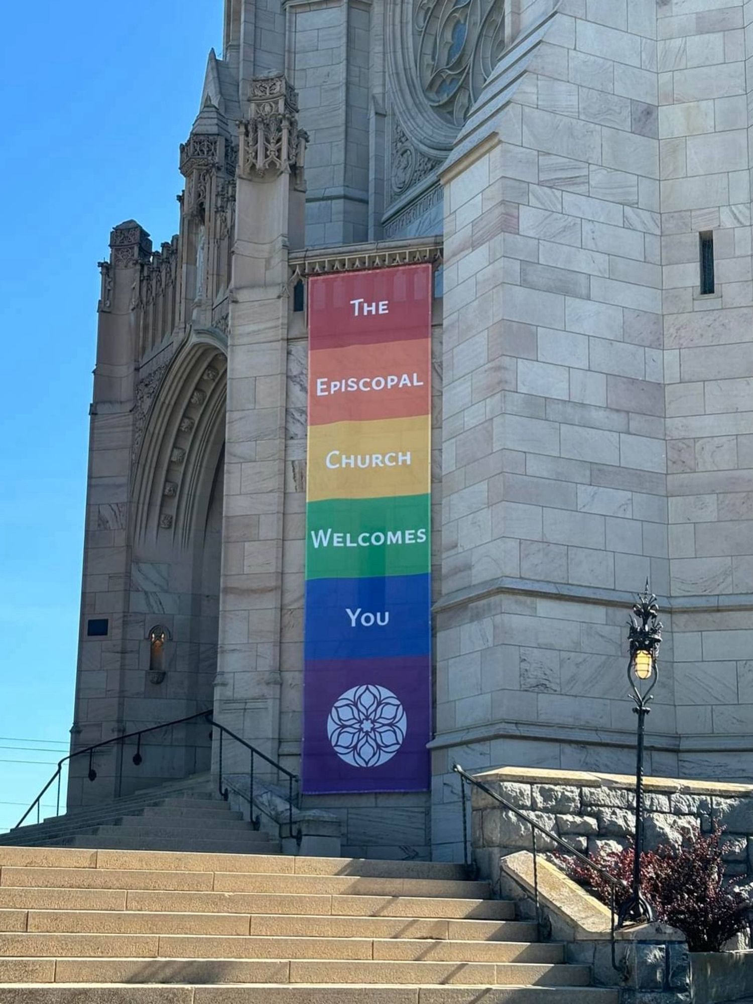 A new banner on my church, the Cathedral of St John the Evangelist, Spokane. Stone steps lead up to the entryway of a neogothic church building. The banner reads The Episcopal Church Welcomes You, with a different color behind each word. At the bottom where the banner is purple is the church logo (a stylization of our rose window)