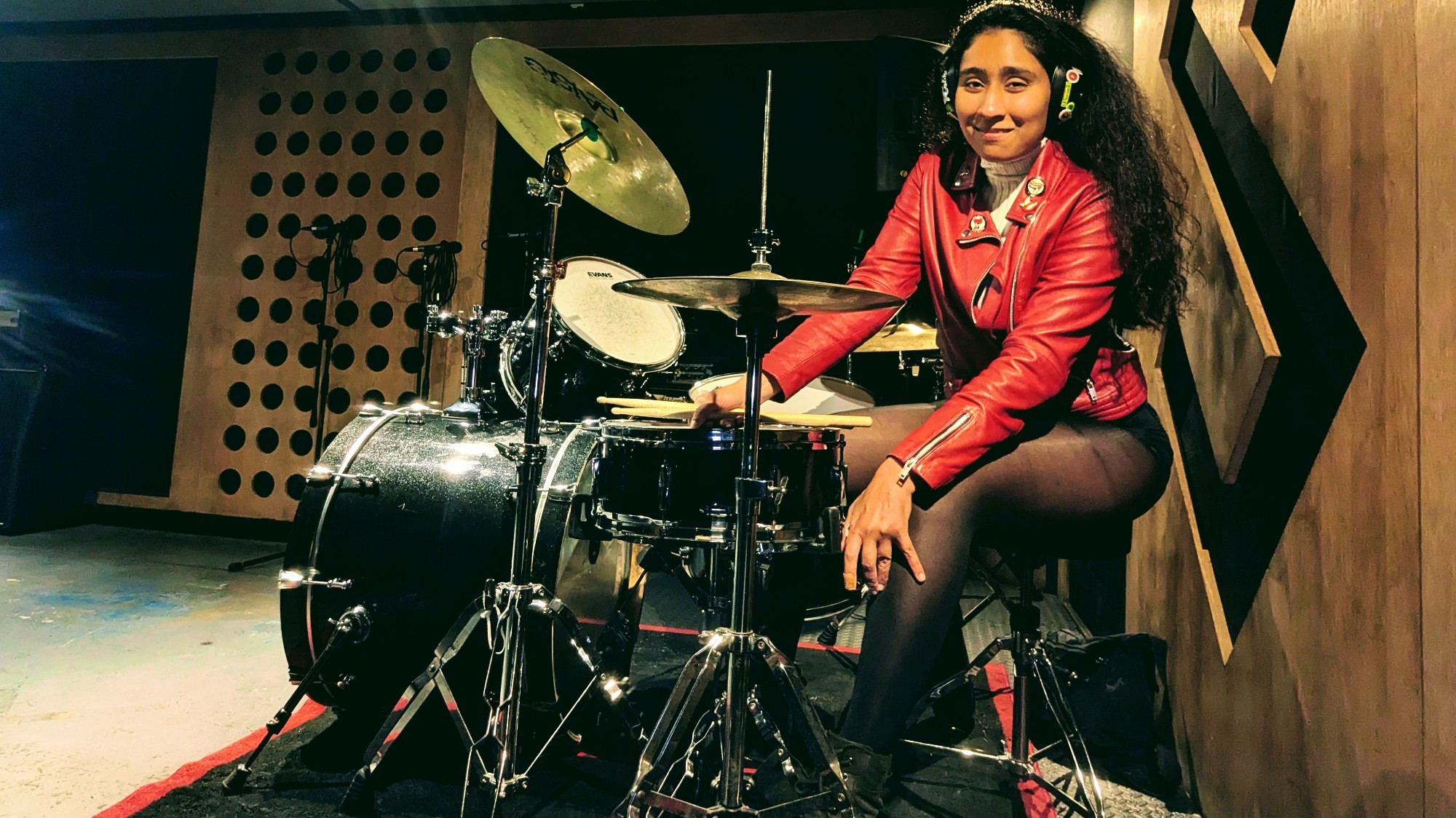 A selfie of me sitting by a black Natal drum kit with a mixture of different cymbals. The Pirate Studios branding is on the wall right behind me. I am wearing a bright red jacket with antifascist badges, a white wooden turtleneck, black hot pants, black tights, and black boots. My long hair is not tied up, it extends to halfway down my back. I am wearing black ear protectors. I am looking towards the camera and smiling while clutching my drumsticks, light coloured Csibi 2A size sticks. I have a handbag on the floor to my right (behind me, from the view of people seeing).