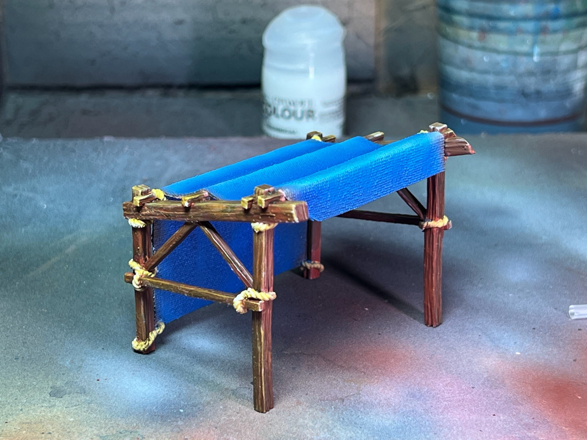 A wooden market stall, with brown beams and a blue tarp and tan ropes. Printed in miniature out of UV reactive resin.