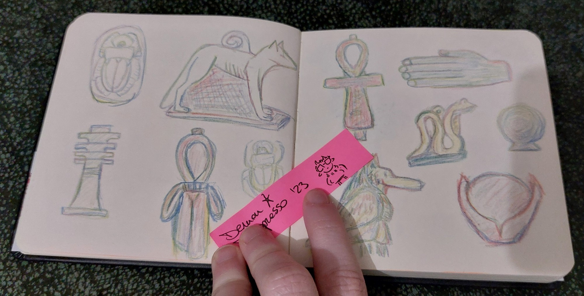 A photo of a square sketbook open flat on a moss green surface. There are sketches of ancient Egyptian amulets done with a rainbow pencil on both pages. At the center there's a bright pink thin sticky note with a little doodle of my sona, and written "demon depresso '23" held there by three of my fingers.