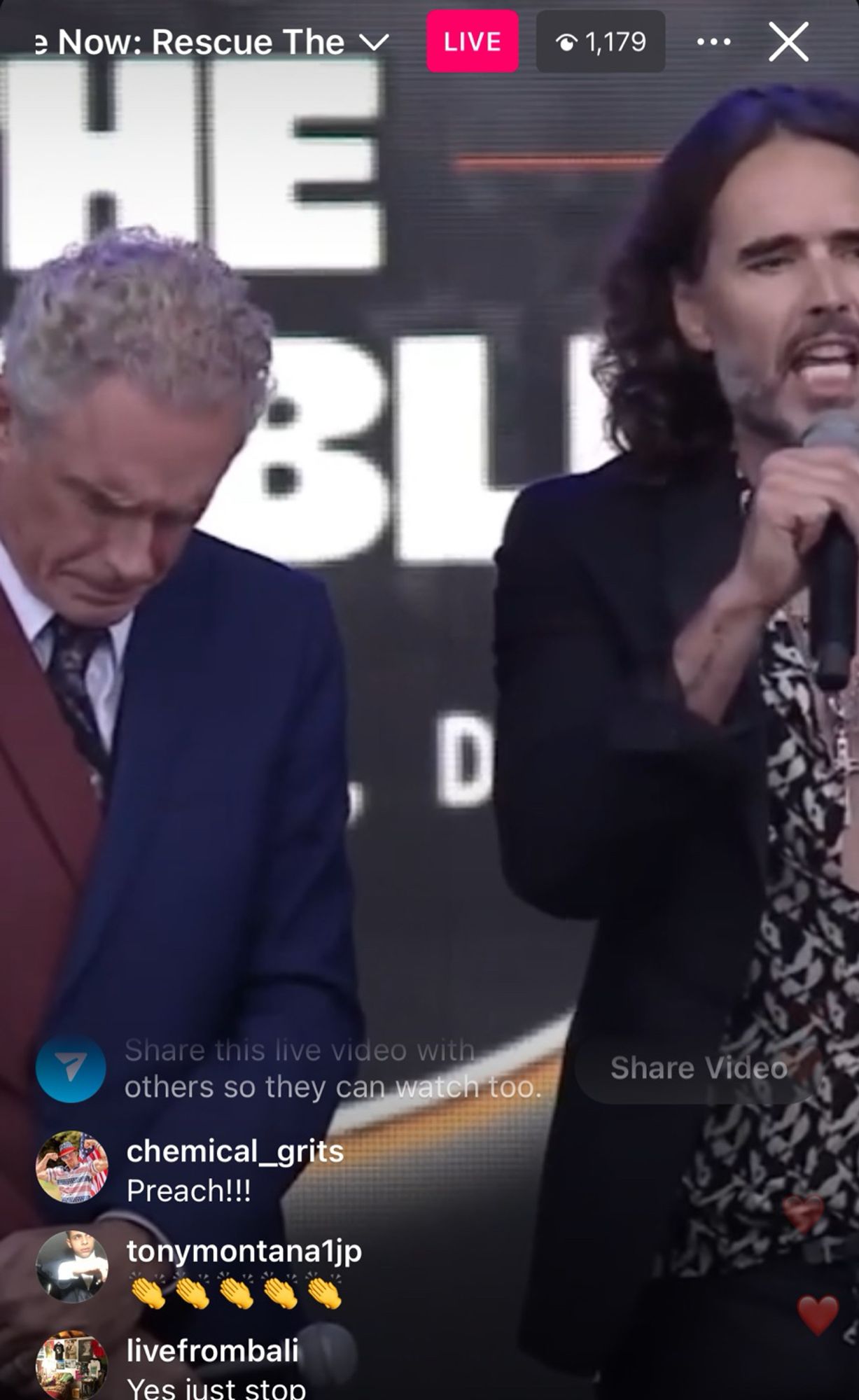 Jordan Peterson in a two-tone suit stands next to Russell brand in a mostly unbuttoned floral shirt and a big gold cross onstage at some horrible fucking  thing