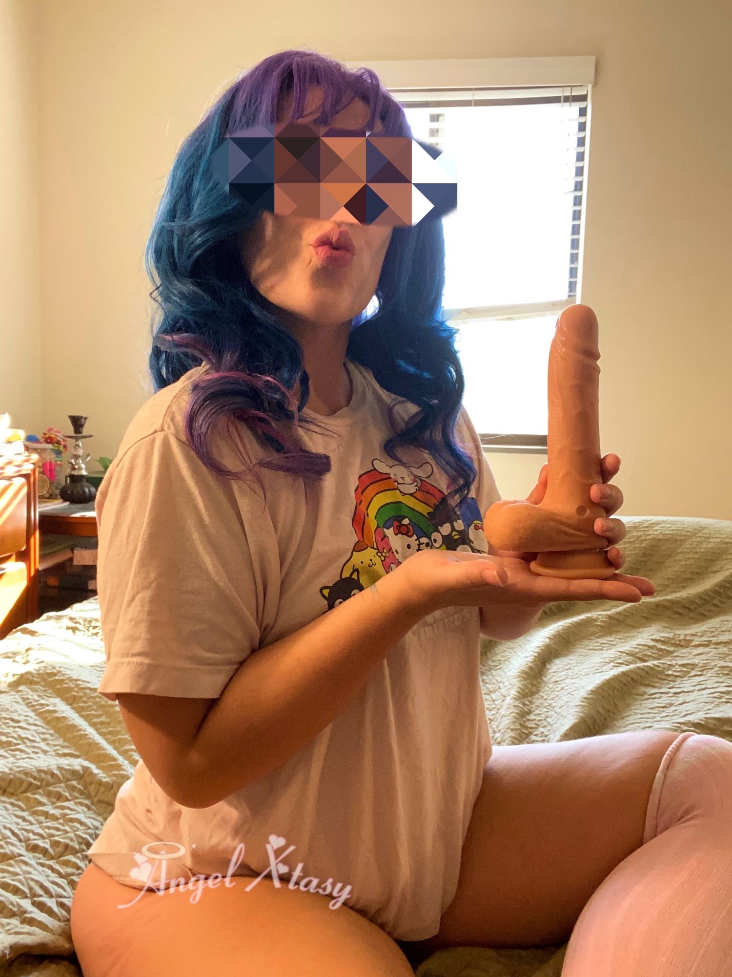 Angel Xtasy, asian transmasc femboy, sitting at the edge of their bed with one leg on and on off. They wear a purple and blue wig, pink sanrio shirt, and pink thigh highs. He holds a dildo up like a trophy. His face is censored