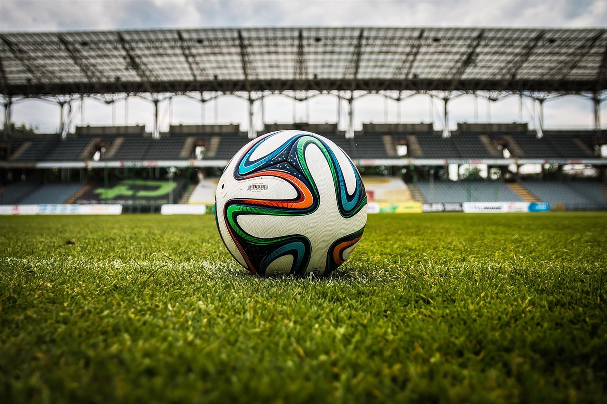 Eine Nahaufnahme eines Fußballs auf einem grünen Spielfeld, mit dem Tor und den Stadionplätzen im Hintergrund unter einem teilweise bewölkten Himmel.