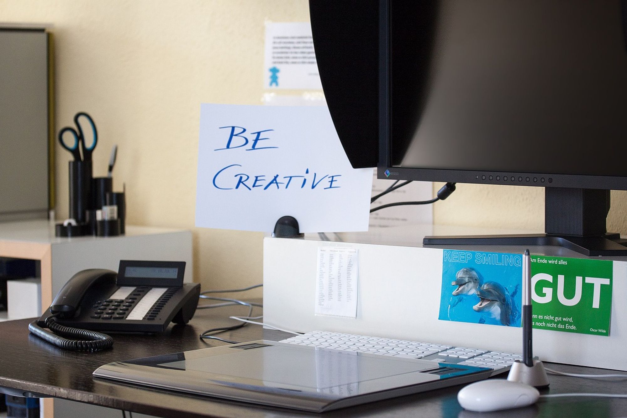 Ein Schreibtisch mit einem Computermonitor, einer Tastatur und einer Maus. Auf dem Tisch steht ein Telefon und ein Stiftehalter. Vor dem Monitor befindet sich ein Schild mit der Aufschrift ‘BE CREATIVE’. Im Hintergrund ist eine Pinnwand mit Notizen zu sehen. Daneben liegt eine Zeitschrift.