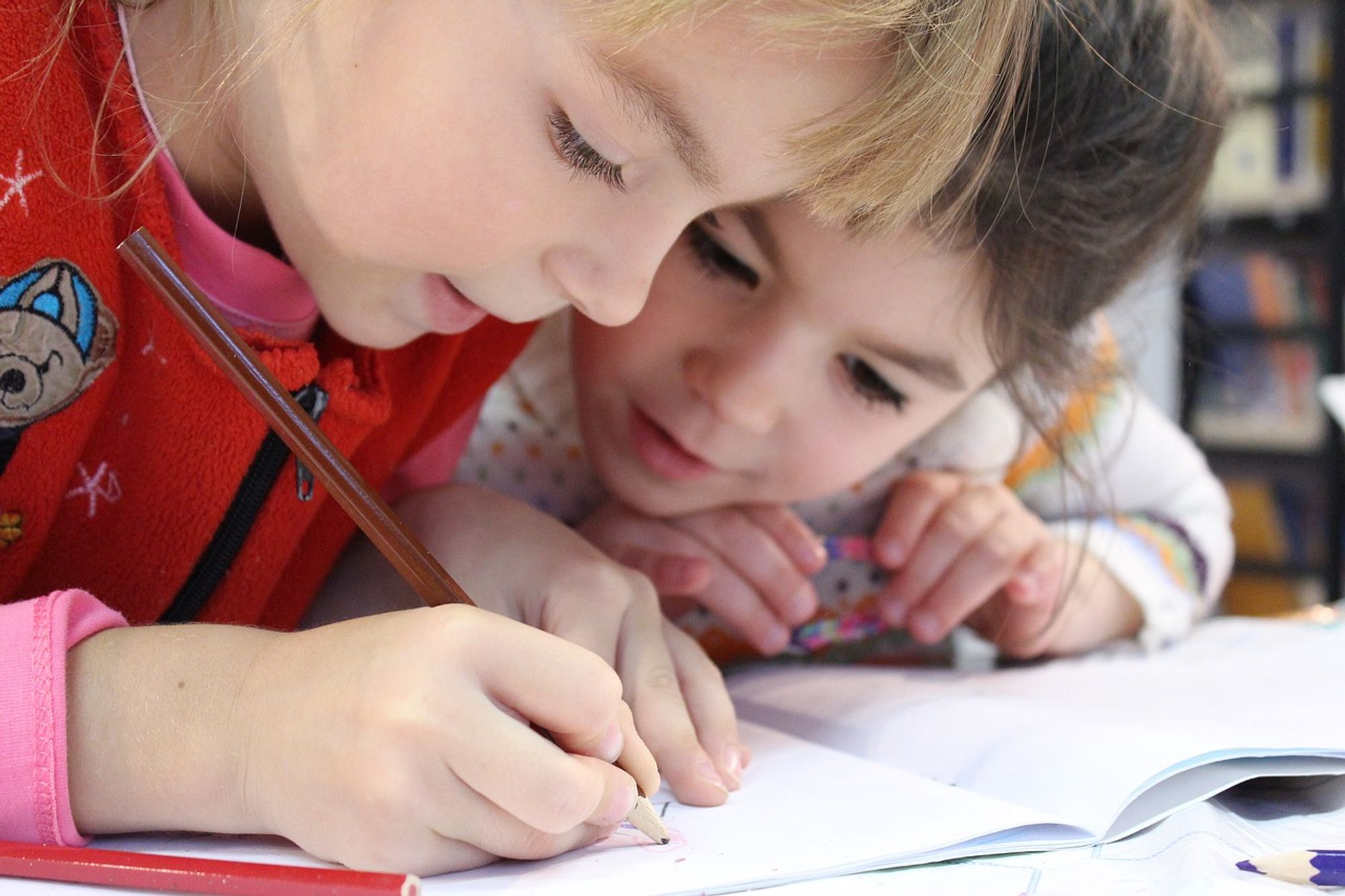 Zwei Kinder sitzen nebeneinander. Ein Kind hält einen Bleistift und schreibt in ein Notizbuch. Der Fokus liegt auf den Händen und der Schreibaktivität, was auf Lernen oder Hausaufgaben hinweist.