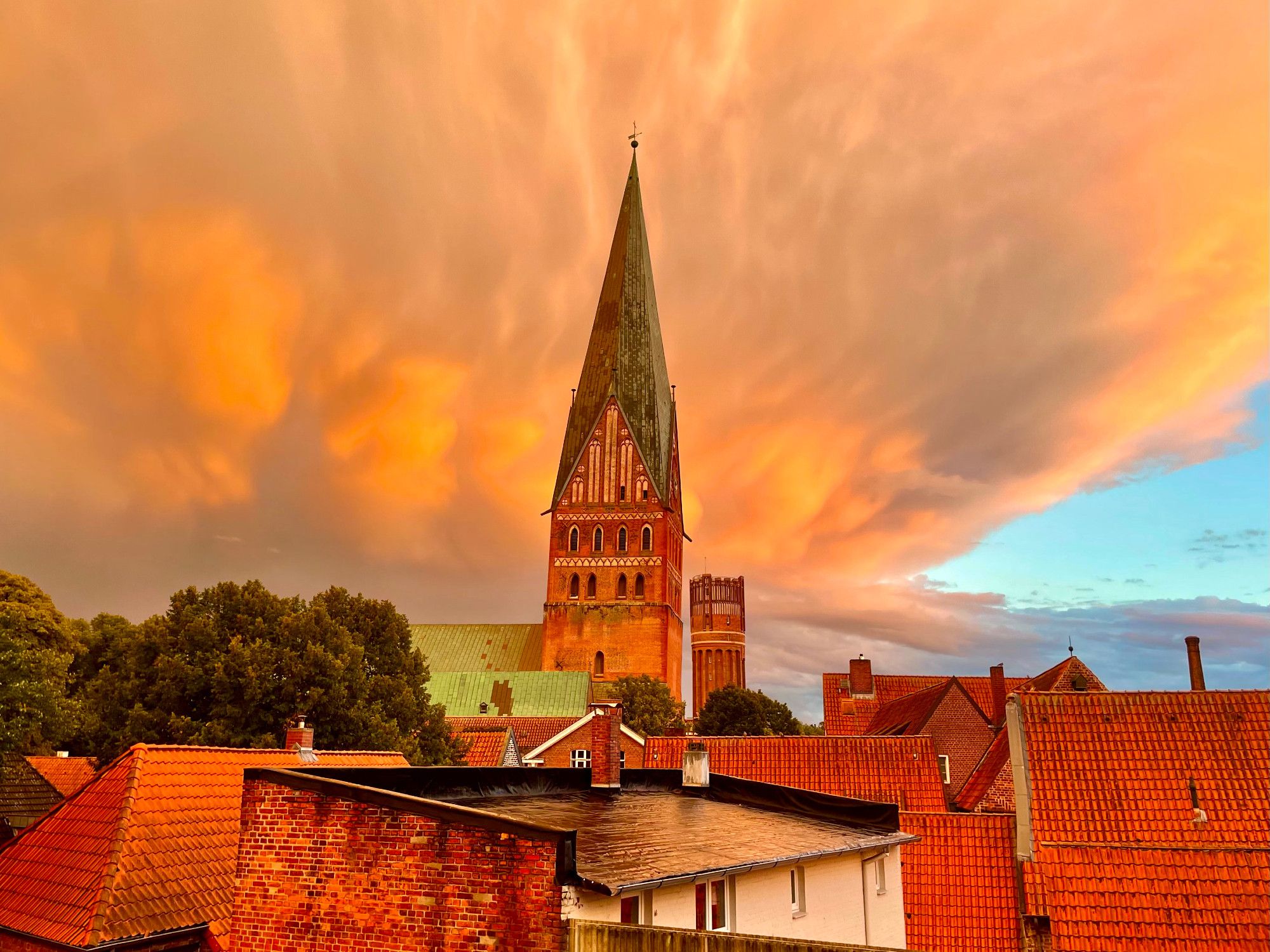 Kirchturm aus Backstein mit kupfergrünem Helm, Dächer, Bäume, Wolken, und all das in ein erstaunliches Abendrot getaucht