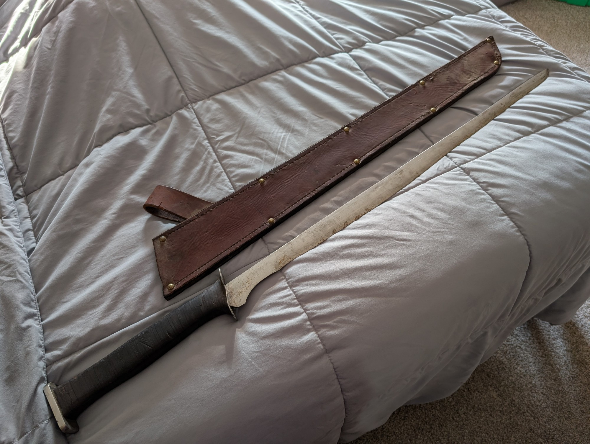 A sword and sheath laying on a bed. The sheath is above the sword. The sheath is brown leather with 10 brass rivets, and a belt loop. The sword is relatively unpolished, with many markings along the blade, which is somewhat reminiscent of a Cutlass. However, the hilt is styled more like you might find on a Chinese sabre, and is wrapped in leather.