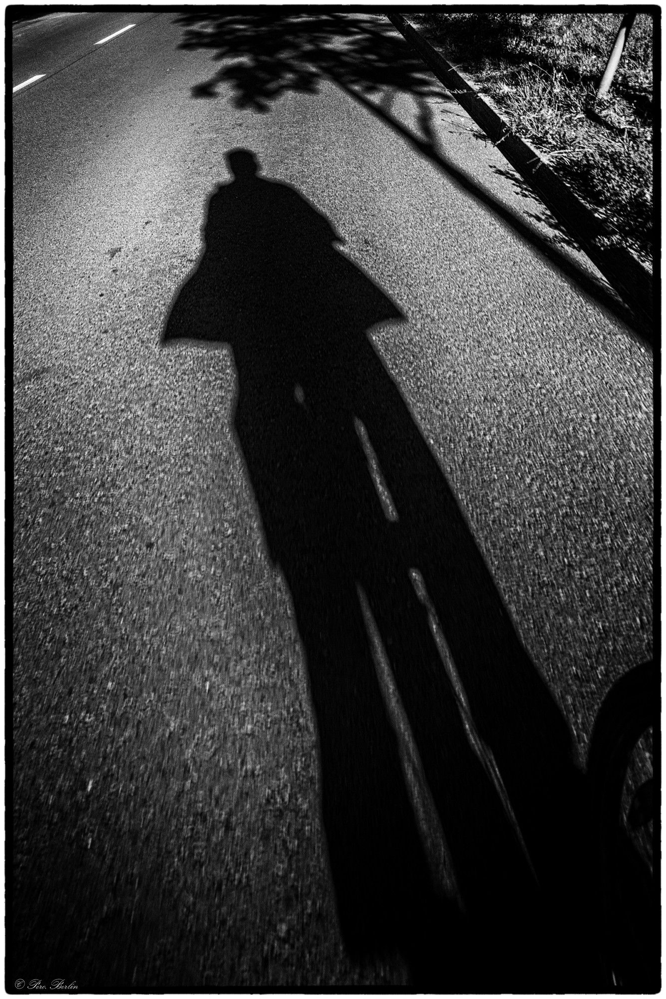 Black and white photograph of the long shadow of a cyclist on the asphalt.