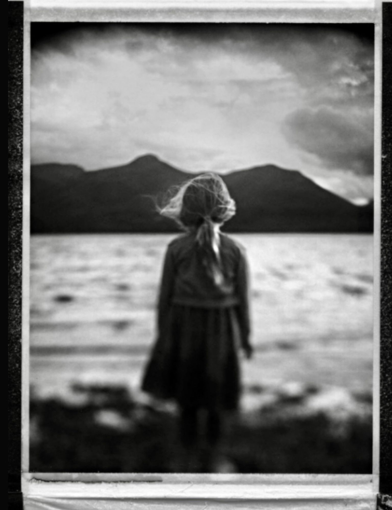 A black and white photograph of a girl standing in front of a lake looking over to a mountain