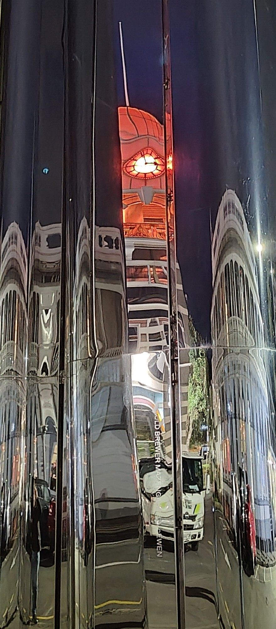 An orange-lit clock tower and surrounding buildings reflected distortedly in the 'funhouse mirror' facade of an art gallery at night.