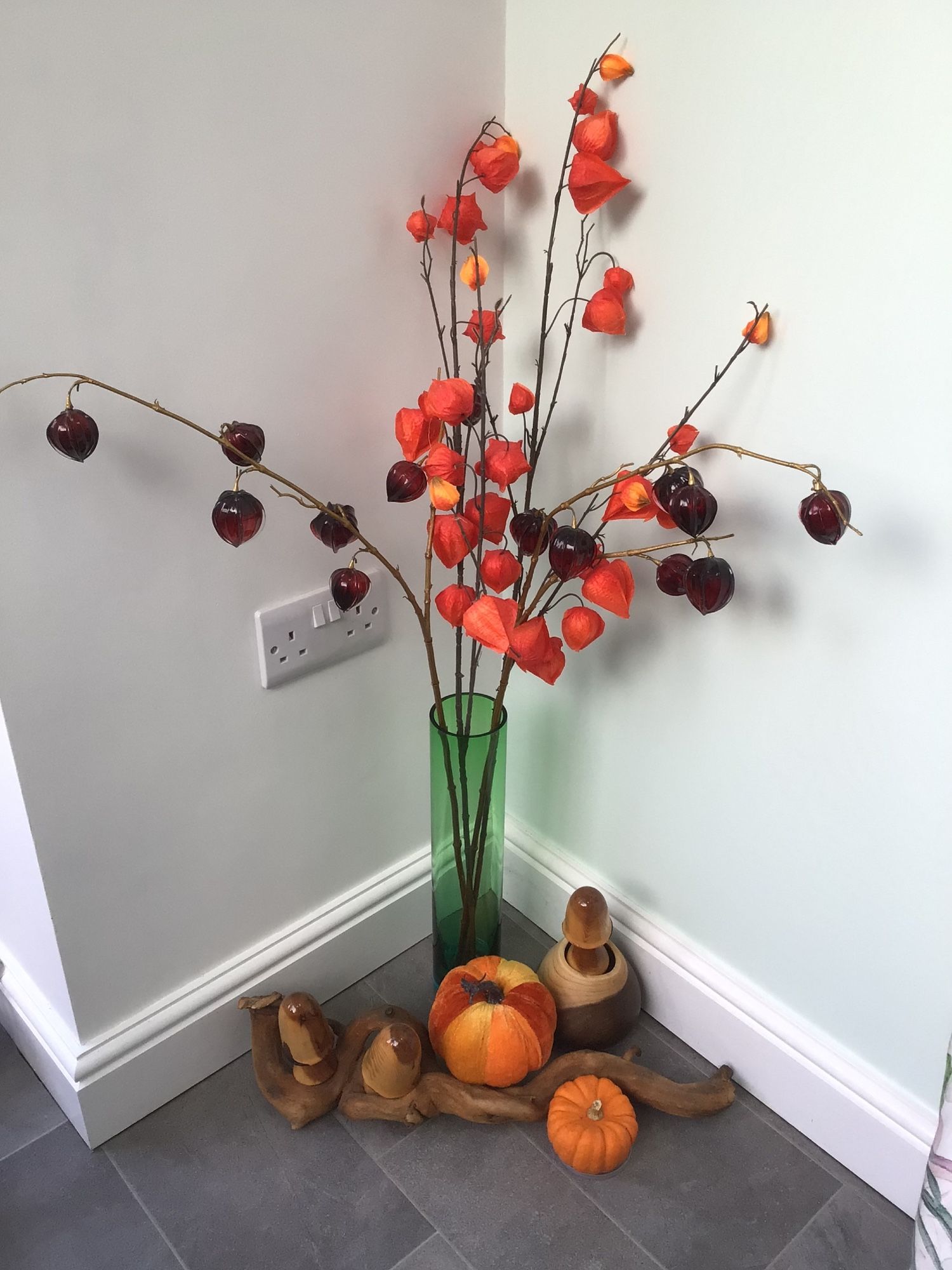 Autumn colour corner-arrangement of artificial Chinese lanterns in a green glass vase, with driftwood, carved wooden toadstools plus real and woven pumpkins.