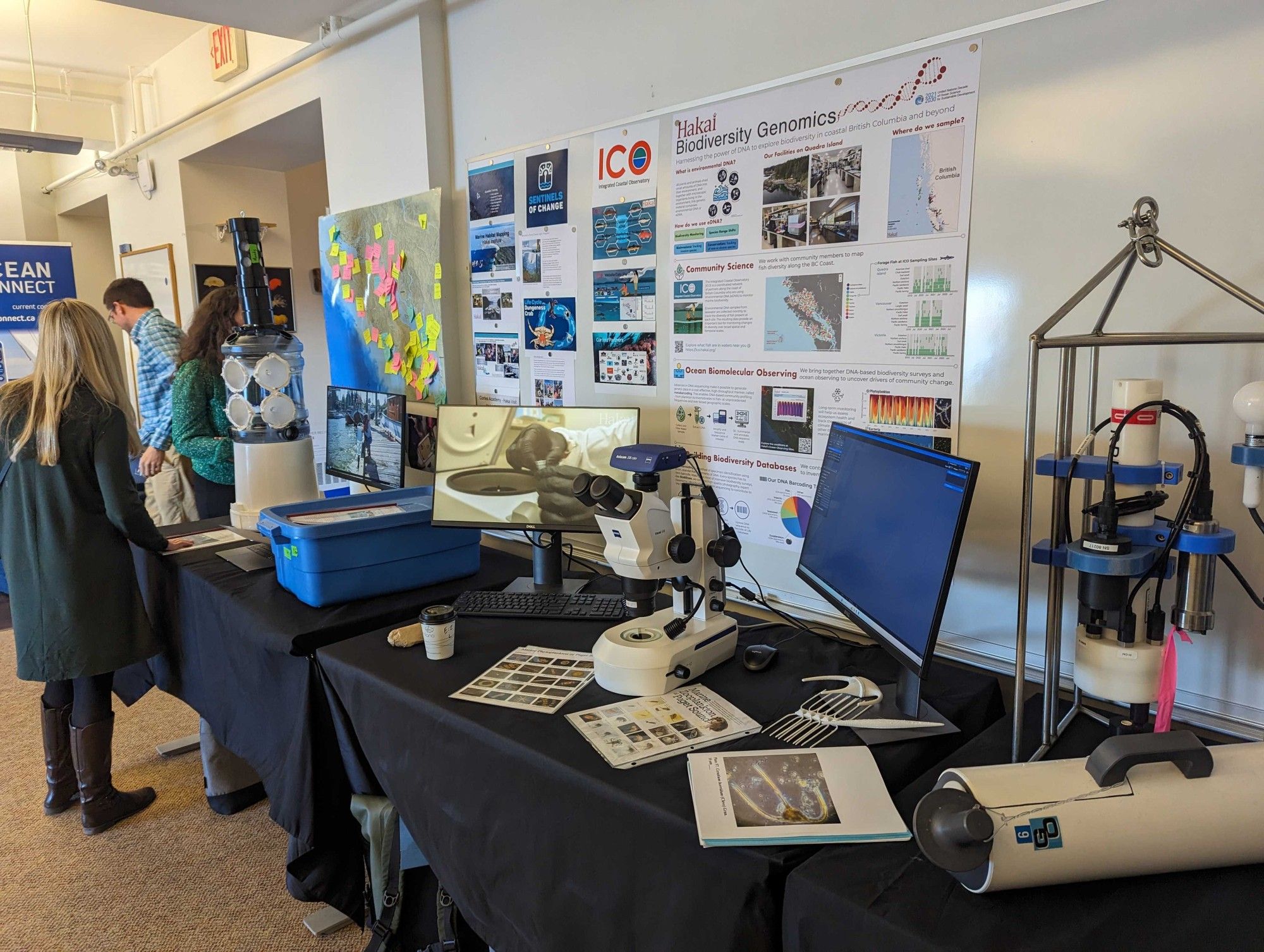 Scientific instrumentation, including a CTD and a microscope set up with a camera and screen, sit on a black table with research posters in the background.