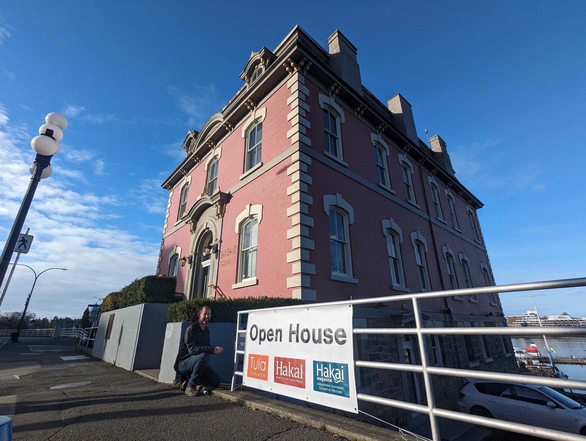 A pinkish-red brick building that houses the Hakai Institute and Tula Foundation stands with a blue sky in the background and an open house sign in front.