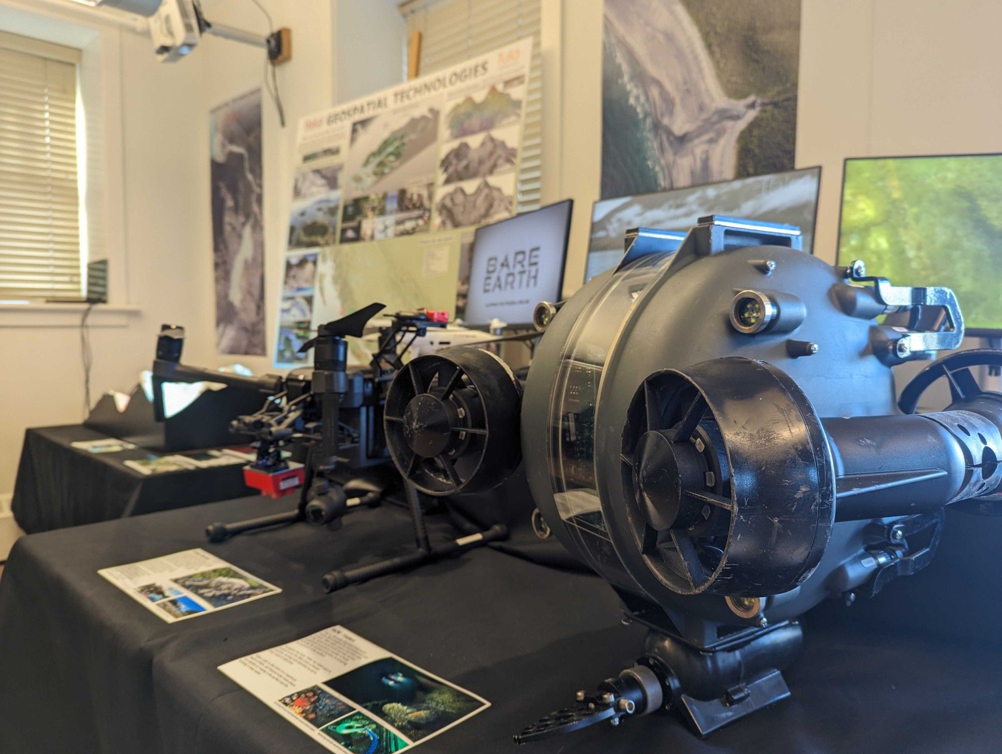 A remotely operated vehicle (ROV) and drones used by Hakai Institute scientists for geospatial research sit on a table, with display posters showing aerial imagery and terrain models in the background.