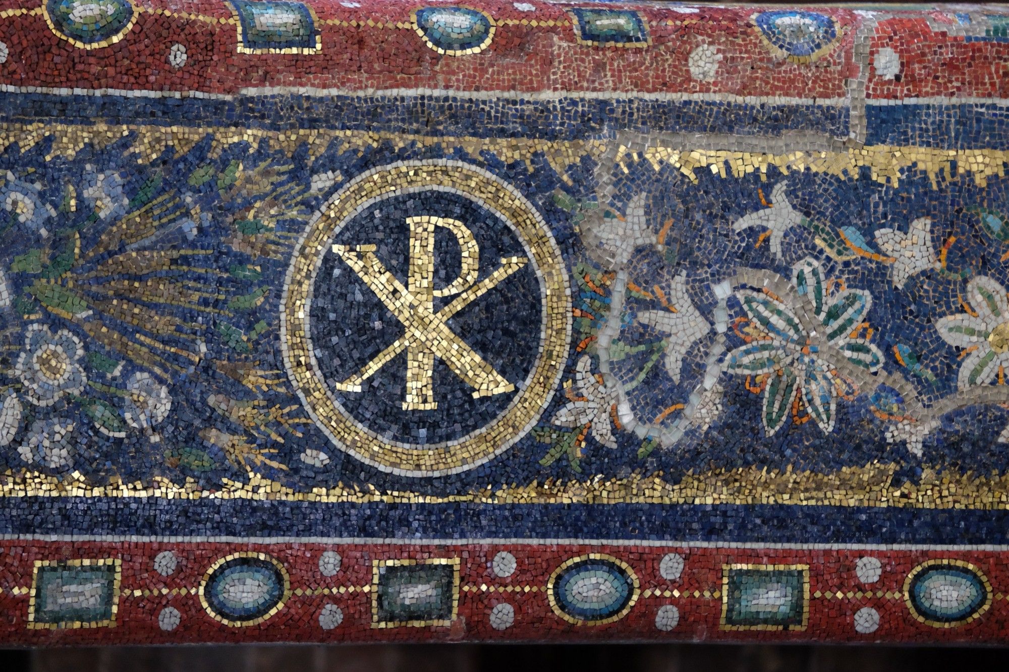 Mosaic featuring a chi-rho symbol enclosed within a circle and surrounded by floral and precious-stone imagery in blue, red, green, white, orange, and gold tesserae.