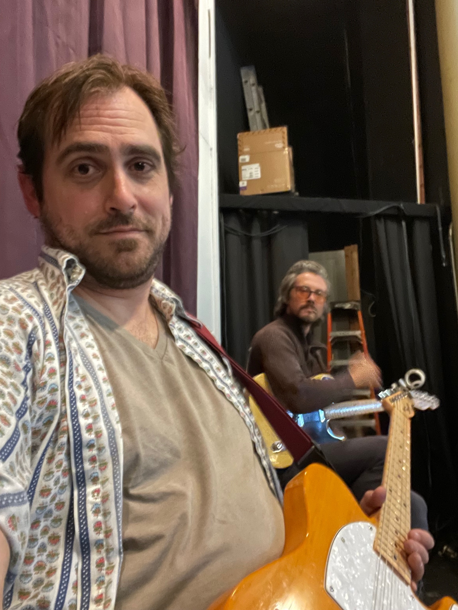 Two fellas holding guitars, with some of the backstage area visible behind the purple and Black curtains