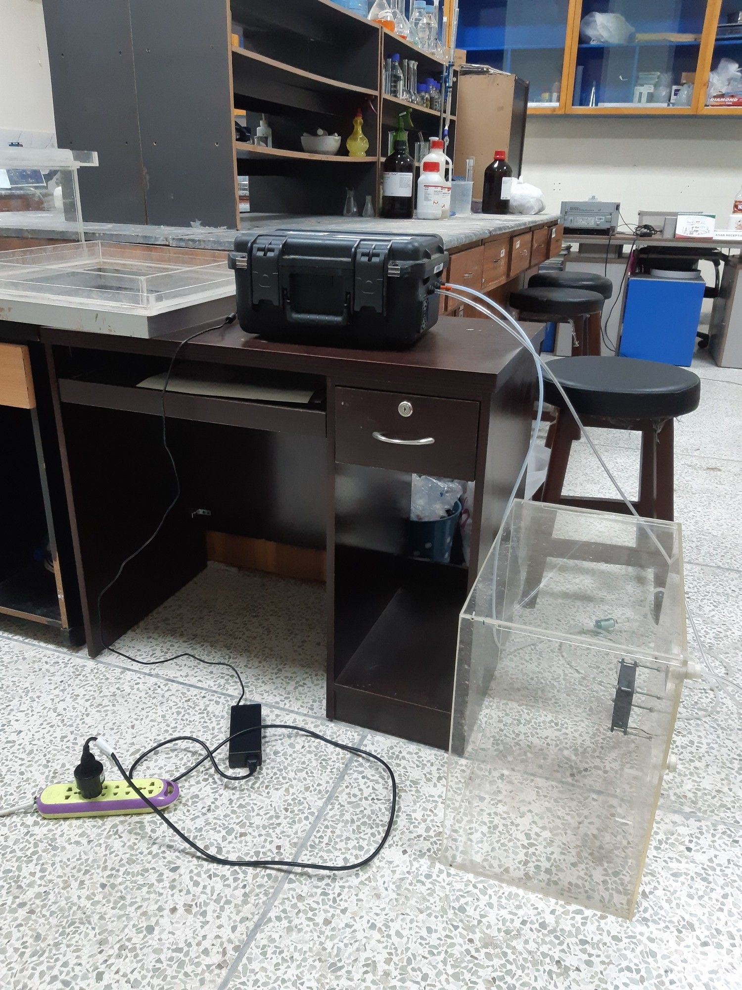 View of a laboratory with lab benches and cupboards, partly made of dark wood. On one table is a black suitcase connected via tubing to a transparent box standing on the floor.