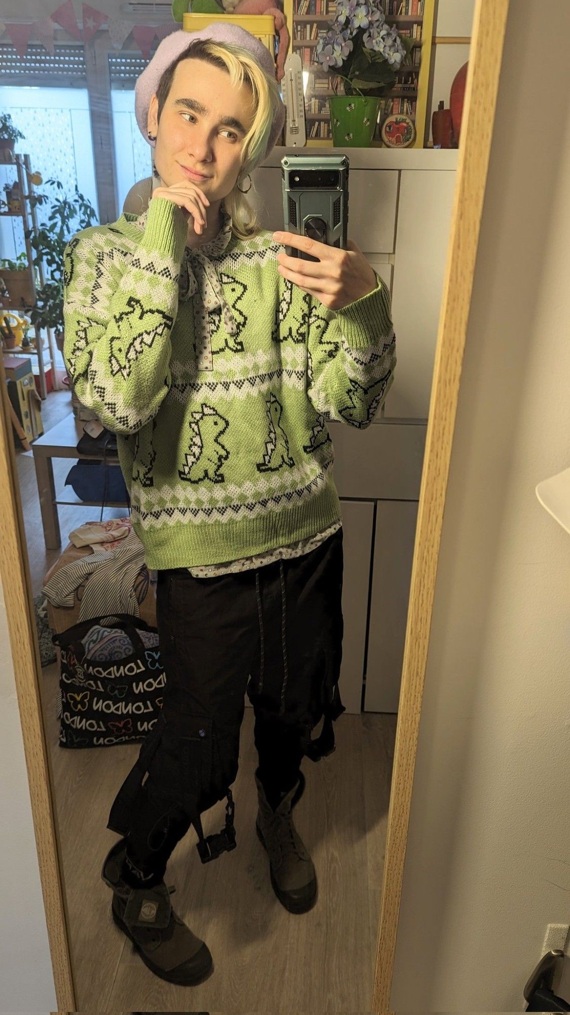 Selfie miroir dans mon appart. J'ai une main devant le menton et je regarde mon portable avec un léger sourire. Je porte un béret Lila pastel, un pull vert tendre avec des motifs façon pull de Noël avec des petits dinosaures, un col lavallière vert d'eau à bois violets et jaunes moutarde dépasse du pull. Je porte également un pantalon cargo noir avec des sangles qui pendentif, des chaussettes noires et des bottines en toile kaki gris.