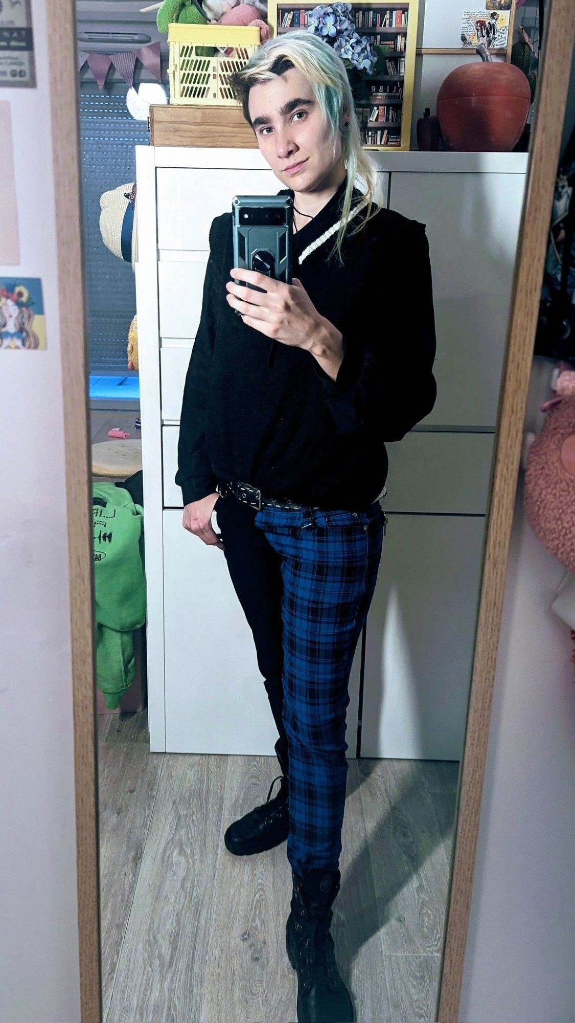 Selfie miroir dans mon appart. J'ai une main dans ma poche de pantalon. Je regarde mon portable. Mes cheveux sont lâchés, on voit bien que le côté long est très dégradé. Je porte une chemise lavallière noire, un pull sans manches noir avec un liseré blanc sur le col en V, un pantalon slim avec une jambe noire et une jambe en tartan bleu et des bottines noires de type rangers mais plus soft.