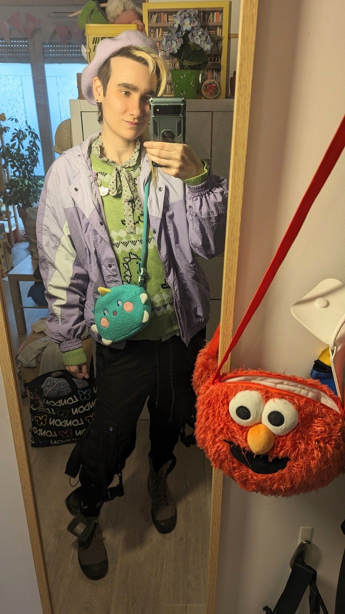 Selfie miroir dans mon appart. Je regarde mon portable avec un léger sourire. Je porte un béret Lila pastel, un blouson de pluie violet pastel, un petit sac peluche verte en forme de petit dinosaure mignon, un pull vert tendre avec des motifs façon pull de Noël avec des petits dinosaures, un col lavallière vert d'eau à bois violets et jaunes moutarde dépasse du pull. Je porte également un pantalon cargo noir avec des sangles qui pendentif, des chaussettes noires et des bottines en toile kaki gris.