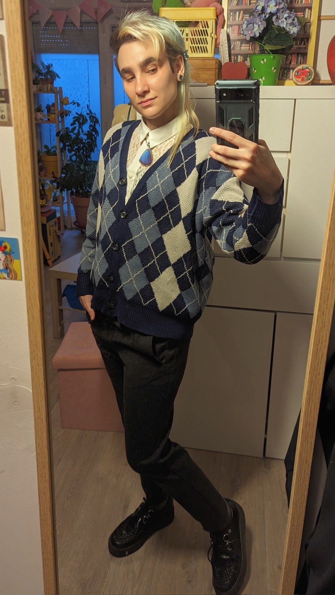 Selfie miroir dans mon appart. J'ai une main dans ma poche de pantalon et je regarde mon portable avec un léger sourire. Mes cheveux sont attachés en demi queue de cheval. Je porte une chemise en dentelle blanc cassé avec un cardigan assez court à motifs jacquard bleu et gris, un pendentif aile de papillon bleu en guise de cravate, un pantalon de costume gris foncé, des chaussettes gris foncé et des creepers noires.