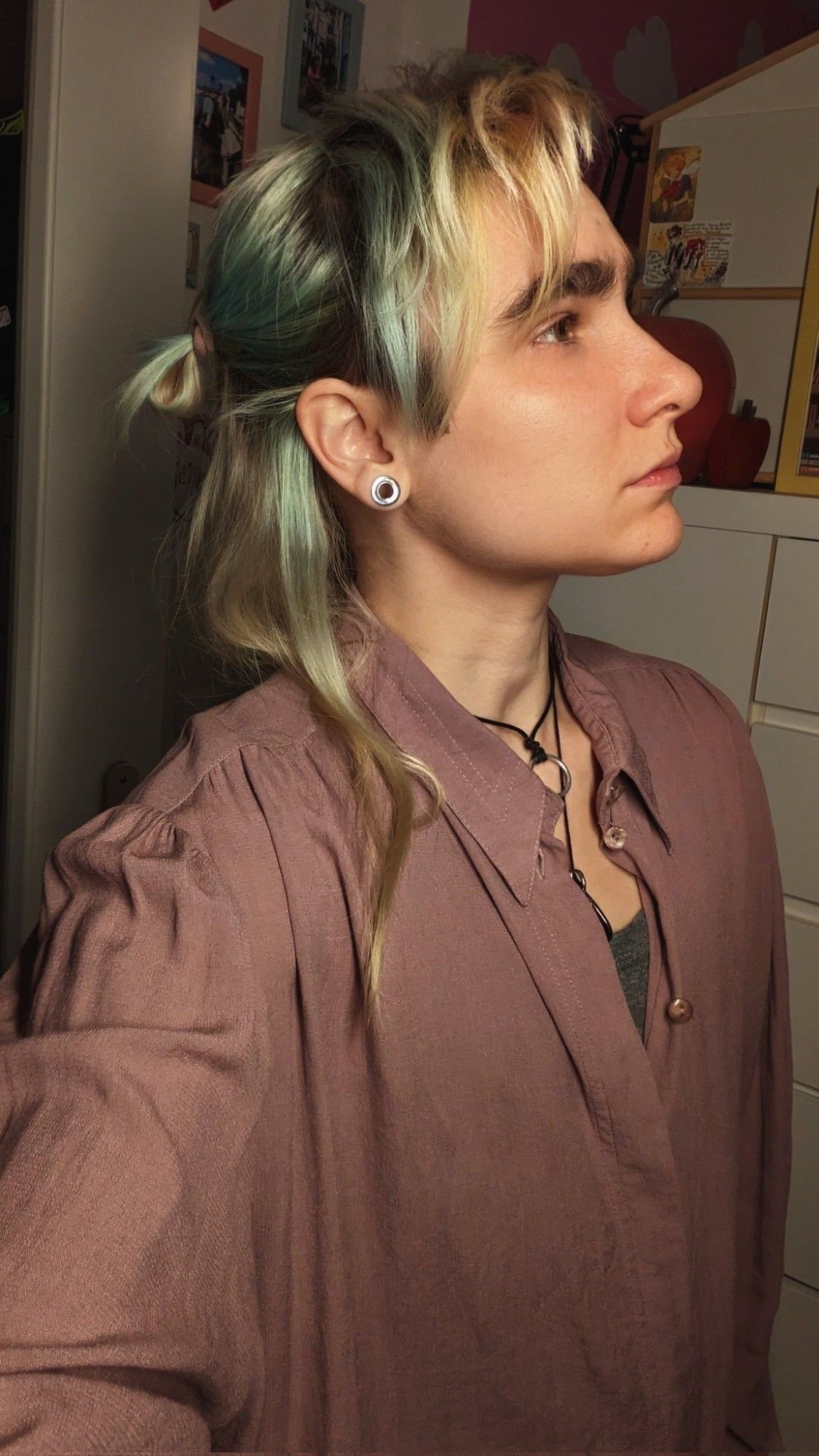 Selfie dans mon appart. Je suis de profil et je regarde devant moi. Mes cheveux sont jaunes et vert pastel, avec une mèche et attachés en demi queue de cheval. Je porte une chemise ample vieux rose portée ouverte avec un trop gris en dessous.