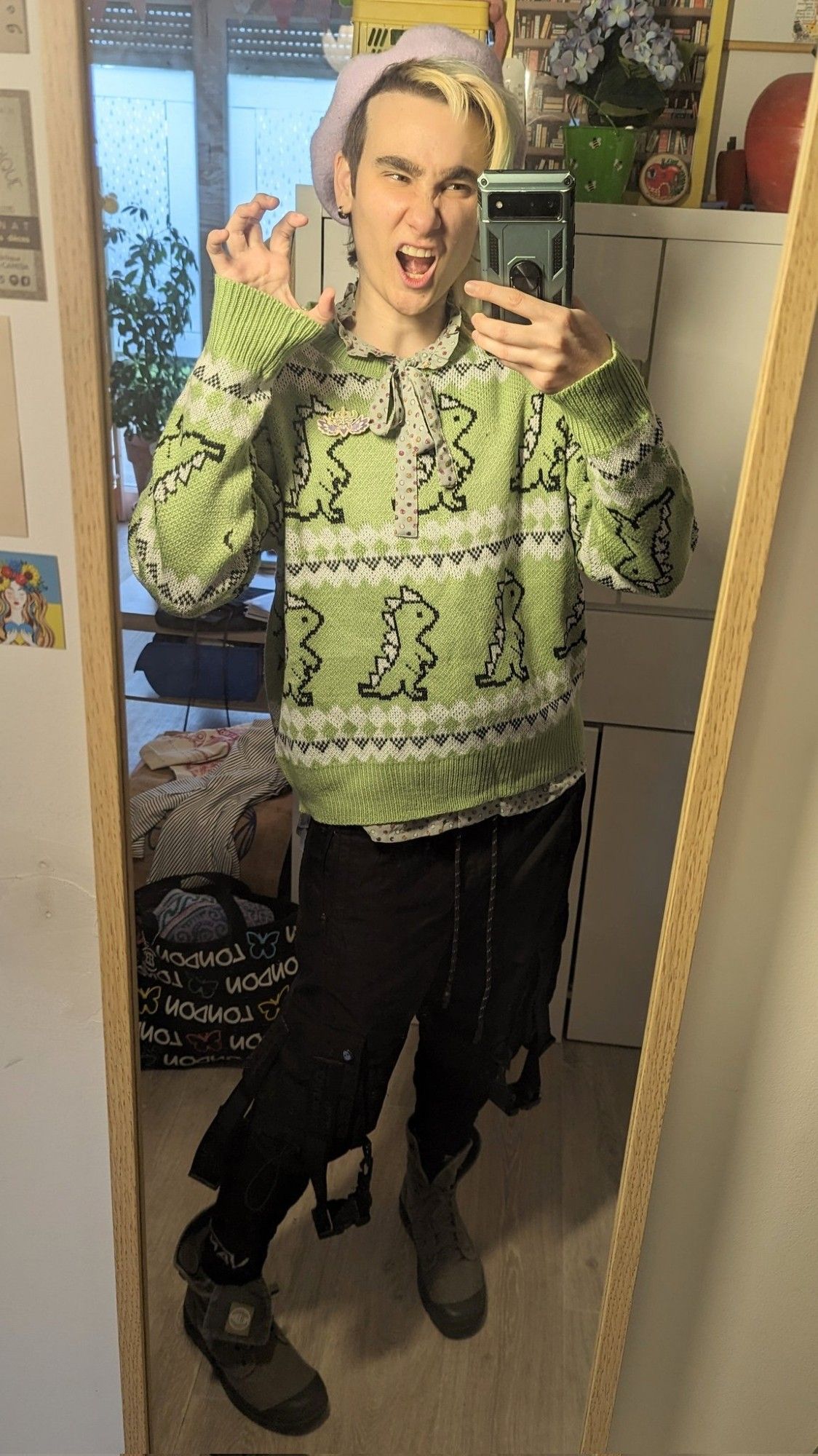 Selfie miroir dans mon appart. J'ai une main crispé pour faire comme si je montrais mes griffes et je fais une grimace comme si je faisais groaaah. Je porte un béret Lila pastel, un pull vert tendre avec des motifs façon pull de Noël avec des petits dinosaures, un col lavallière vert d'eau à bois violets et jaunes moutarde dépasse du pull. Je porte également un pantalon cargo noir avec des sangles qui pendentif, des chaussettes noires et des bottines en toile kaki gris.