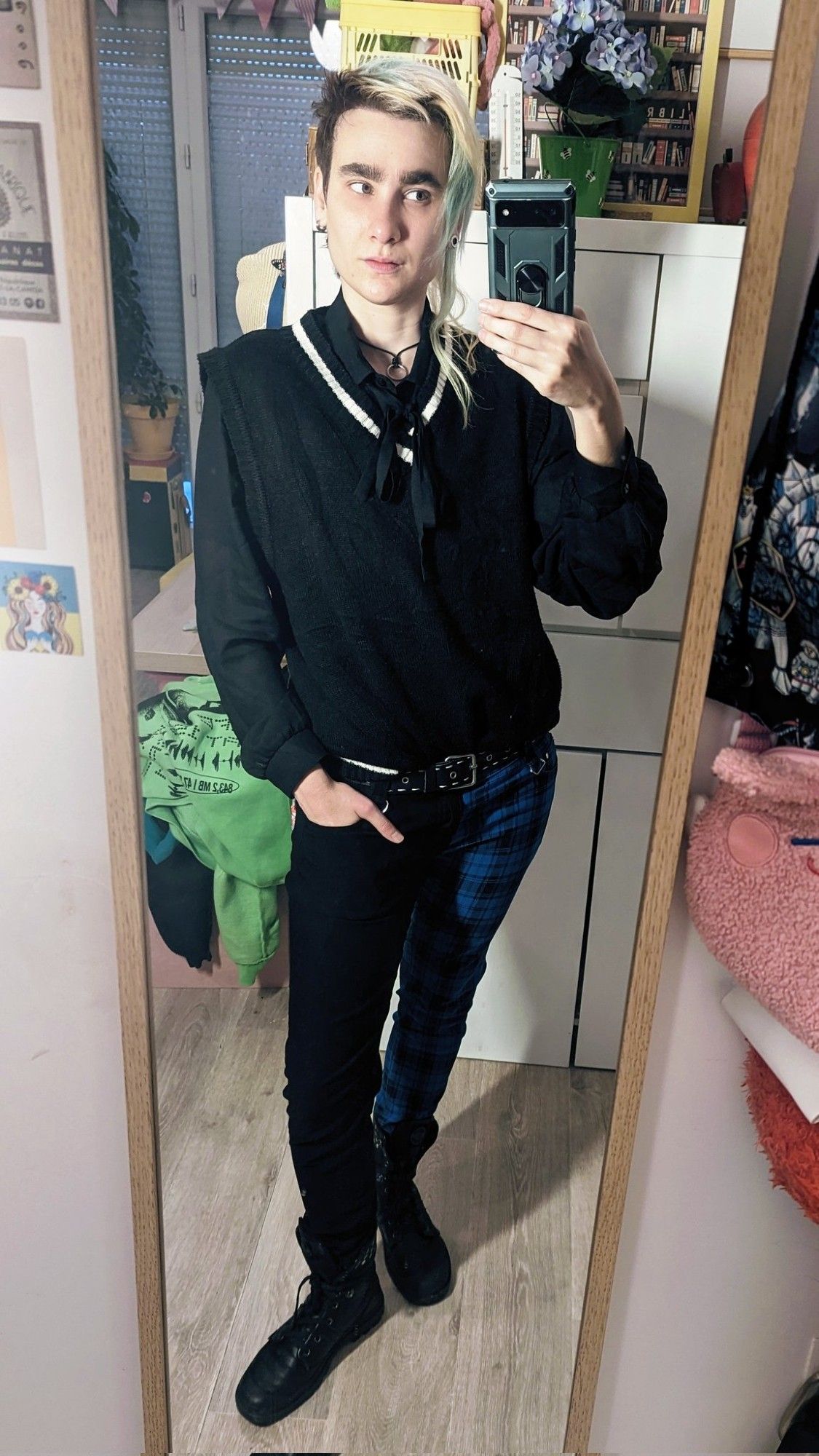 Selfie miroir dans mon appart. J'ai une main dans ma poche de pantalon. Je regarde mon portable. Mes cheveux sont lâchés, on voit bien que le côté long est très dégradé. Je porte une chemise lavallière noire, un pull sans manches noir avec un liseré blanc sur le col en V, un pantalon slim avec une jambe noire et une jambe en tartan bleu et des bottines noires de type rangers mais plus soft.