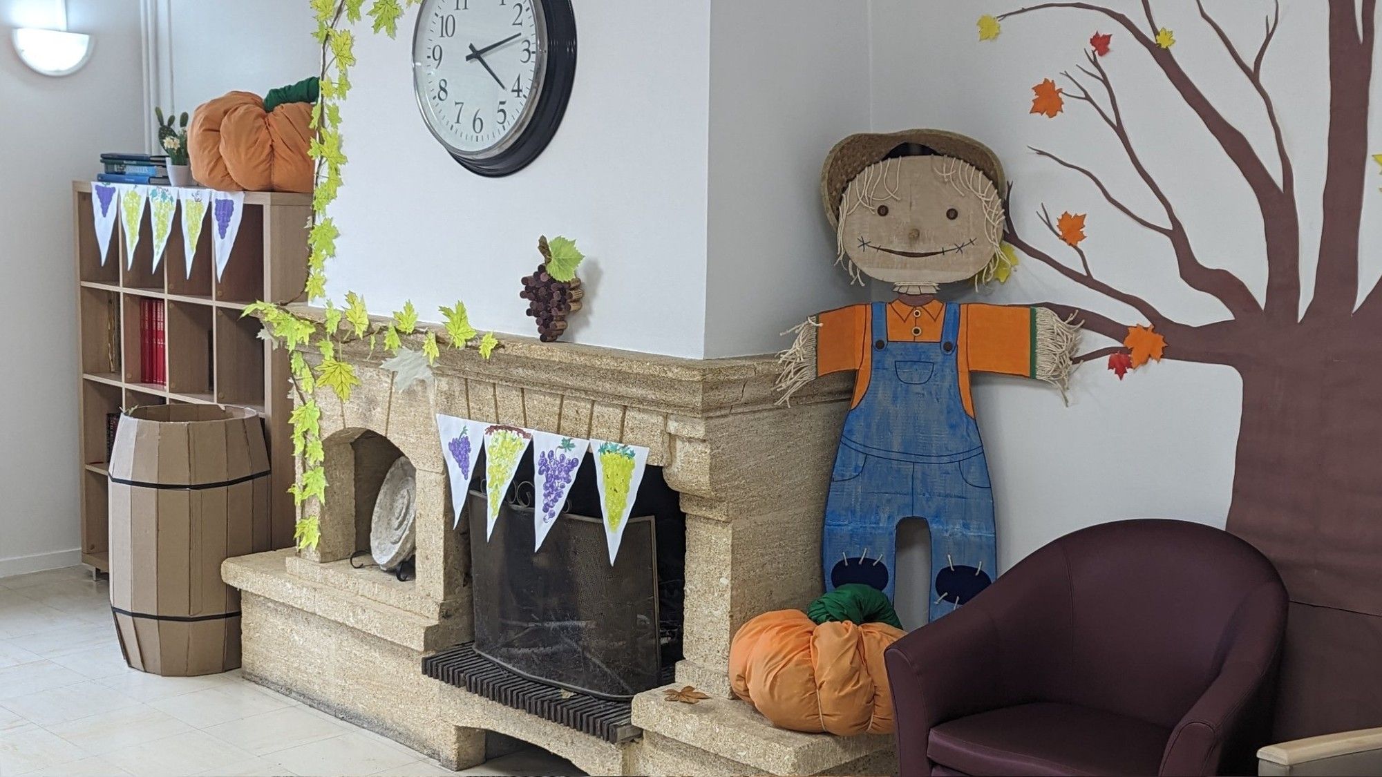 Photo d'un salon avec une déco d'automne diy. Il y a un arbre en carton sur un mur, l'arbre a des feuilles orangées. A côté de l'arbre, contre la cheminée, il y a un épouvantail rigolo fait en carton. A ses pieds il y a une grosse citrouille faite en tissu.
Un tonneau en carton est posé entre une cheminée et une étagère. Sur l'étagère il y a une fausse branche de vigne et des fausses feuilles de vigne sur le tonneau.